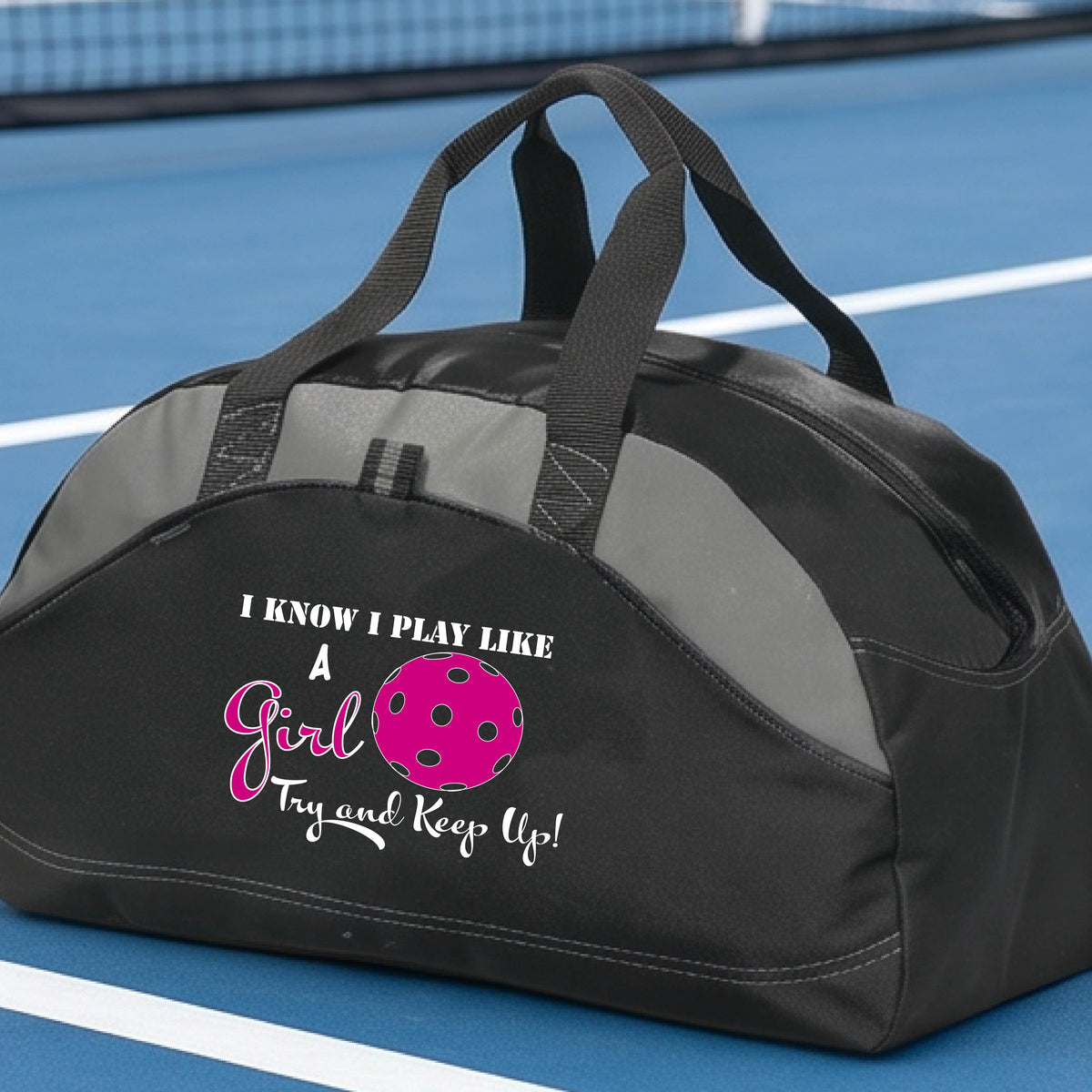 A close-up of a black and gray medium size duffel bag with a white graphic of ‘I KNOW I PLAY LIKE A GIRL TRY AND KEEP UP’ in white and pink with a pink pickleball icon, in front of a pickleball court. 
