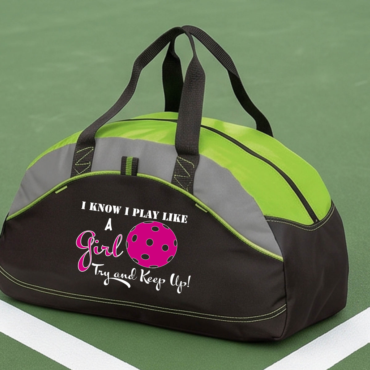 A close-up of a lime green, black and gray medium size duffel bag with a white graphic of ‘I KNOW I PLAY LIKE A GIRL TRY AND KEEP UP’ in white and pink with a pink pickleball icon, in front of a pickleball cour