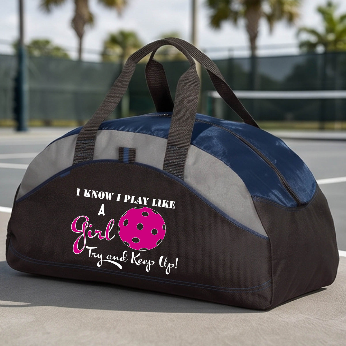 A close-up of a navy blue, black and gray medium size duffel bag with a white graphic of ‘I KNOW I PLAY LIKE A GIRL TRY AND KEEP UP’ in white and pink with a pink pickleball icon, in front of a pickleball cour