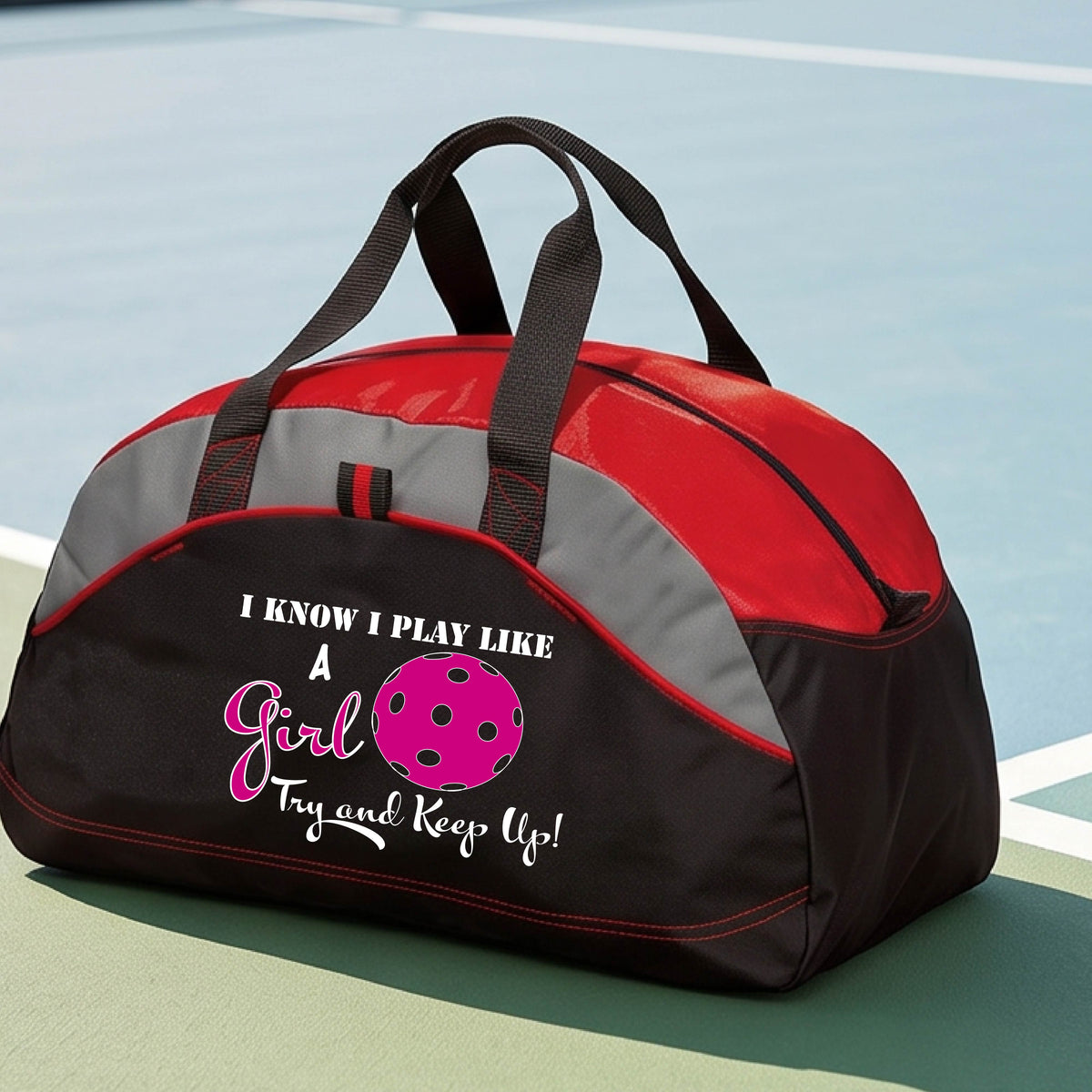A close-up of a red, black and gray medium size duffel bag with a white graphic of ‘I KNOW I PLAY LIKE A GIRL TRY AND KEEP UP’ in white and pink with a pink pickleball icon, in front of a pickleball cour