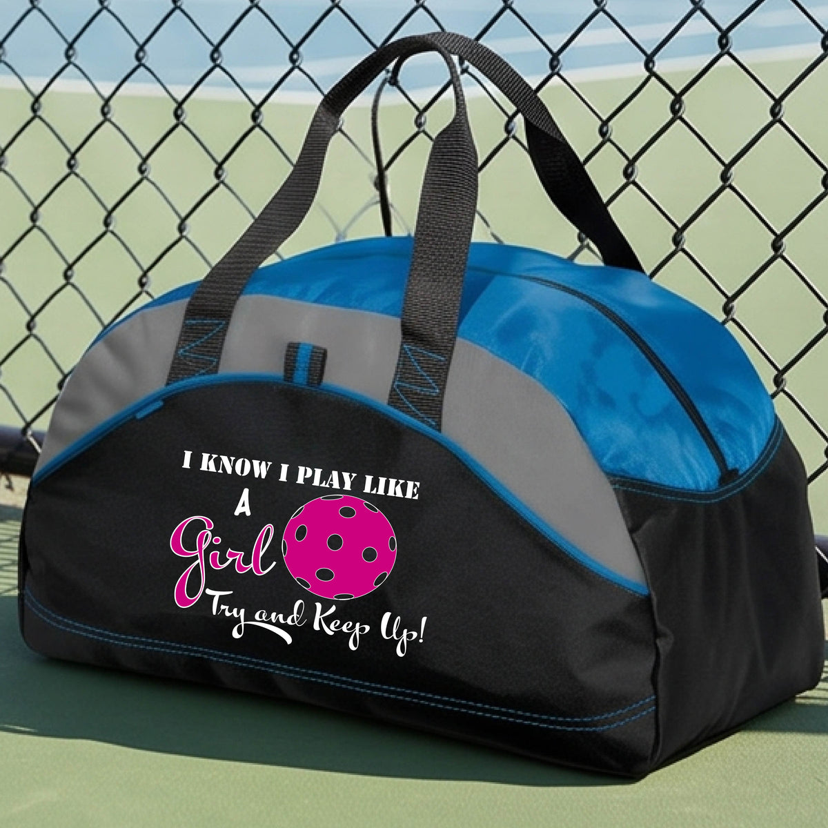 A close-up of a royal blue, black and gray medium size duffel bag with a white graphic of ‘I KNOW I PLAY LIKE A GIRL TRY AND KEEP UP’ in white and pink with a pink pickleball icon, in front of a pickleball cour