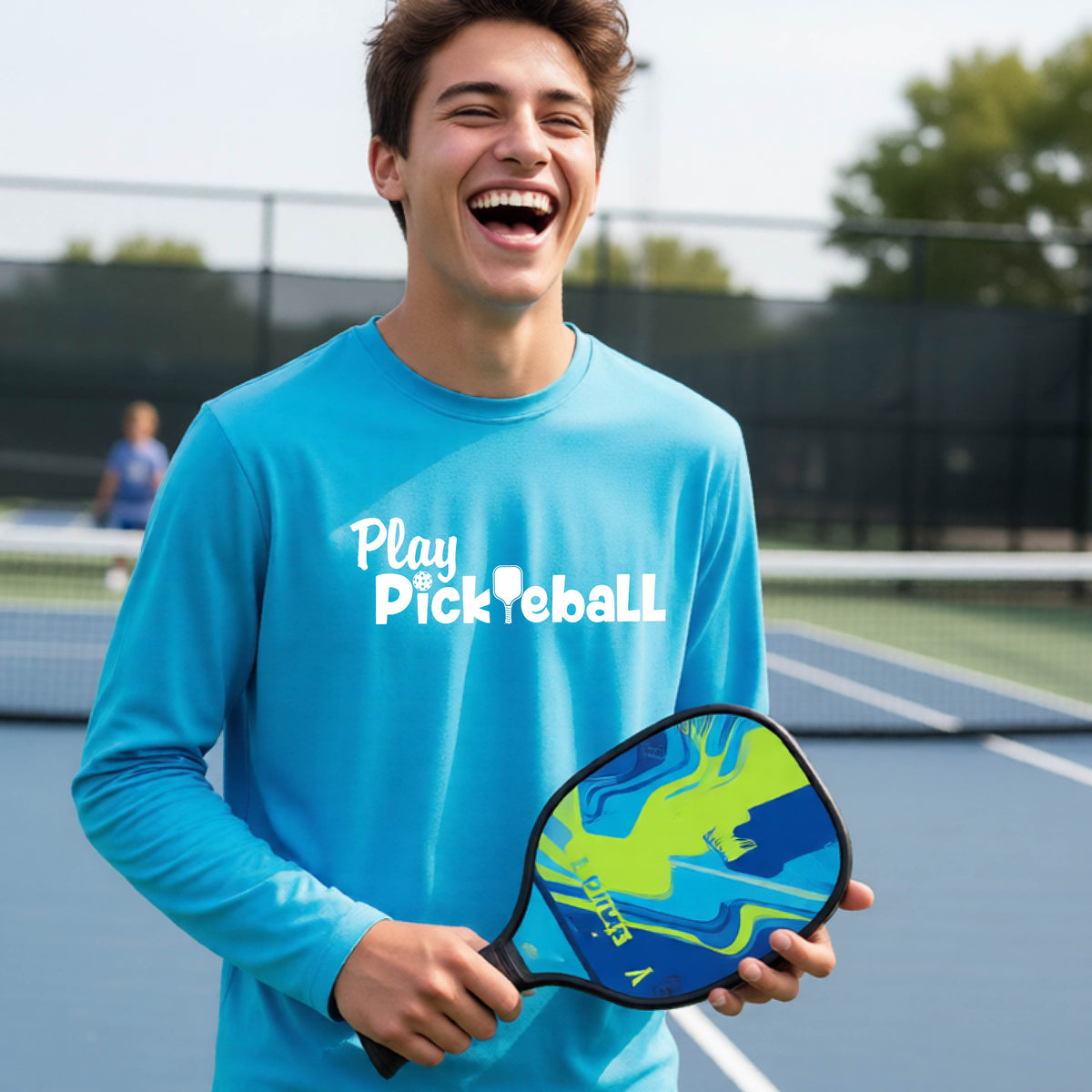 A young person on a pickleball court wearing a bright blue long-sleeve t-shirt with a white graphic of the text 'PLAY PICKLEBALL' across the front.