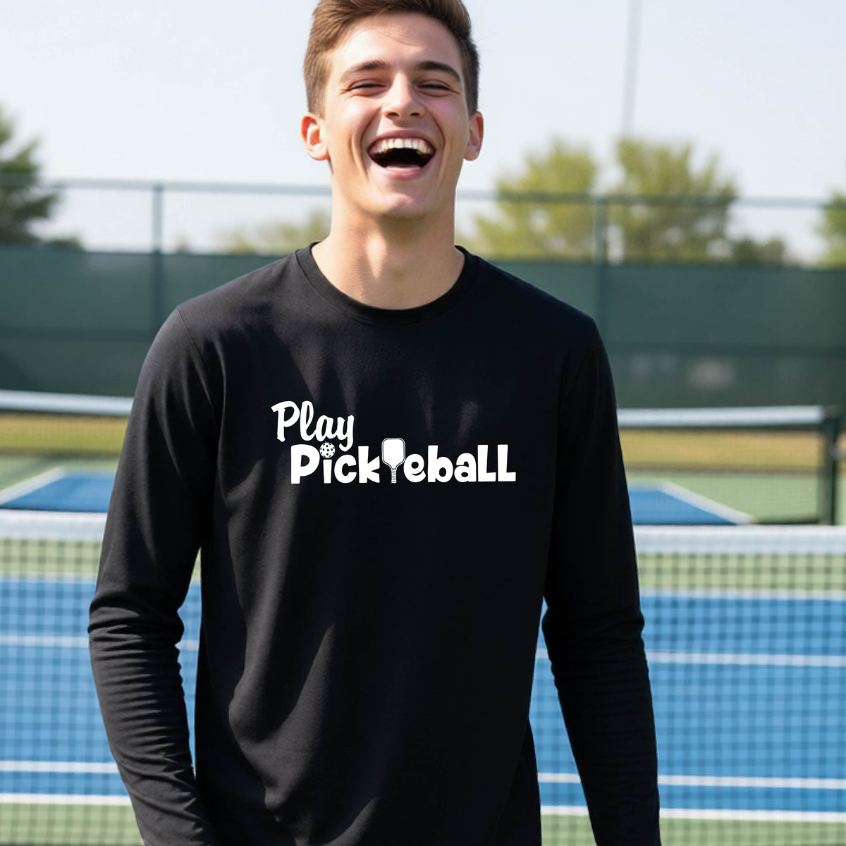 A young person on a pickleball court wearing a black long-sleeve t-shirt with a white graphic of the text 'PLAY PICKLEBALL' across the front.