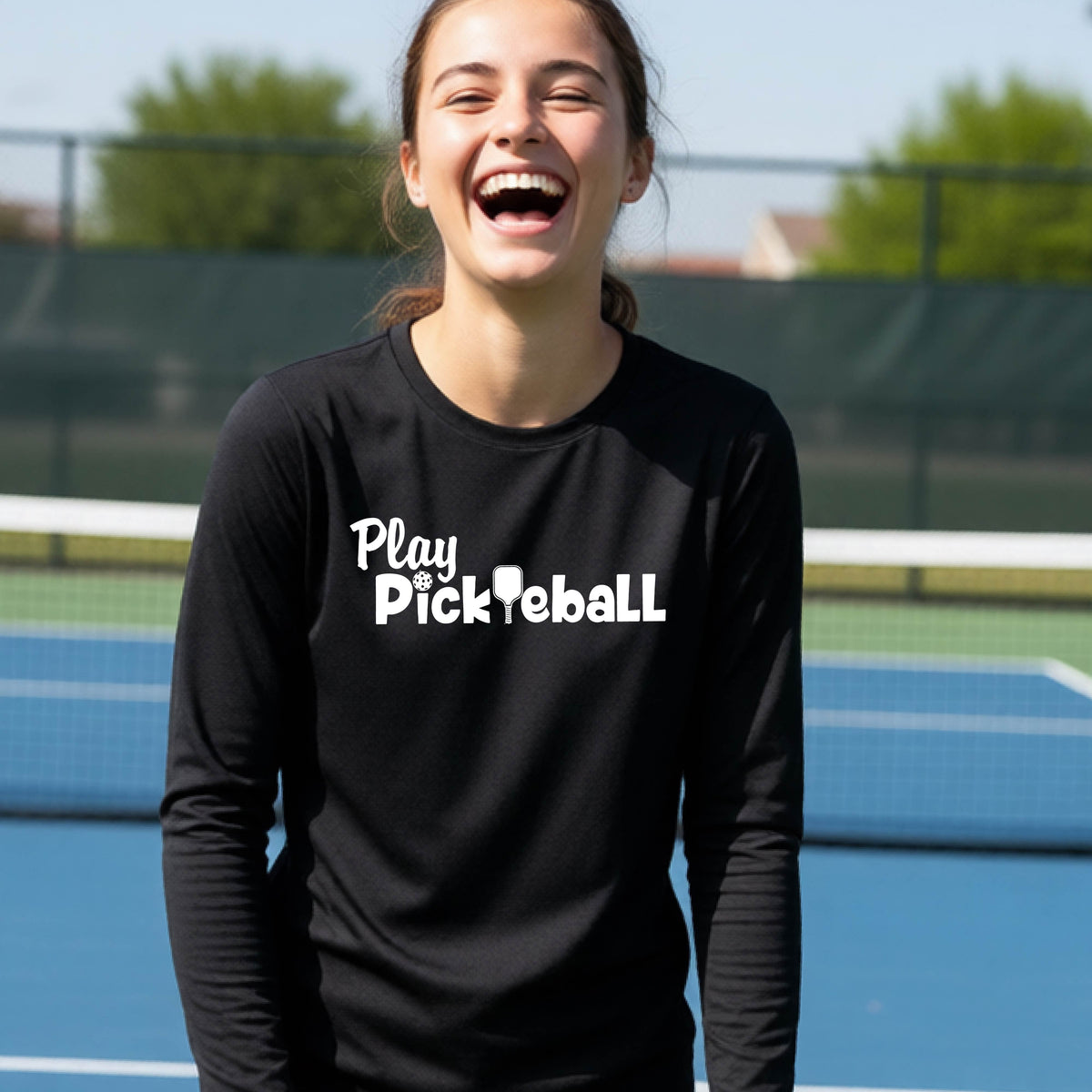 A young person on a pickleball court wearing a black long-sleeve t-shirt with a white graphic of the text 'PLAY PICKLEBALL' across the front.