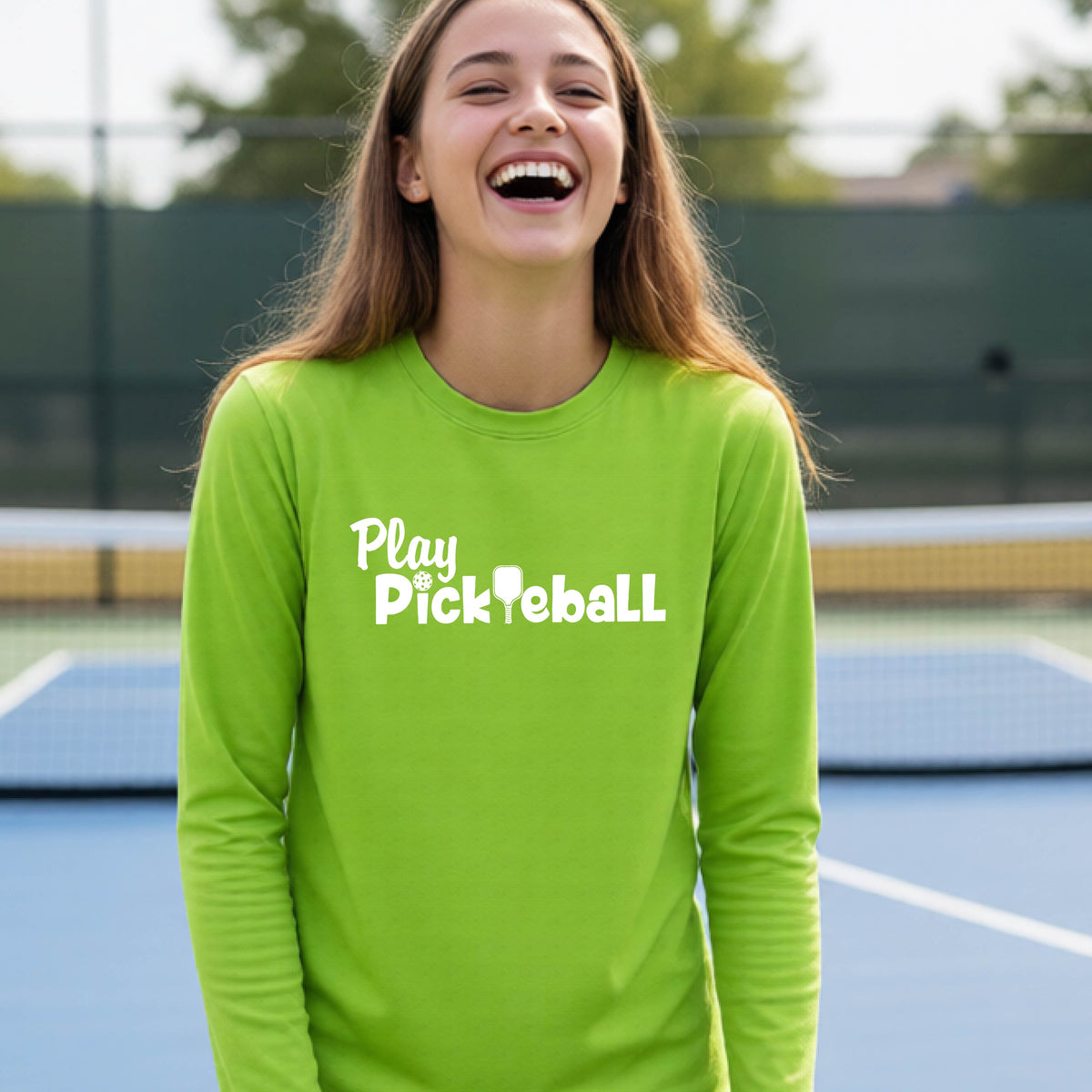 A young person on a pickleball court wearing a lime green long-sleeve t-shirt with a white graphic of the text 'PLAY PICKLEBALL' across the front.