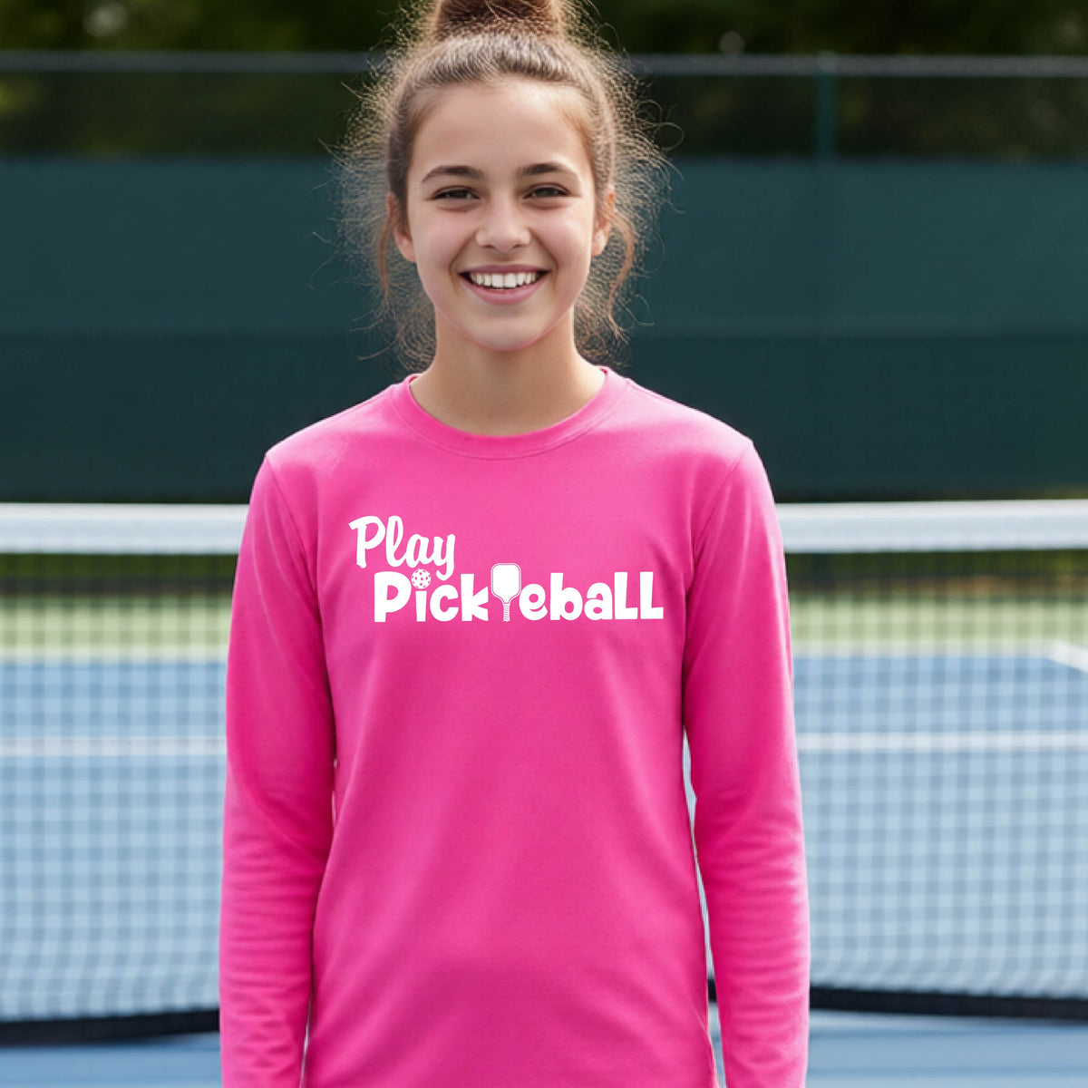 A young person on a pickleball court wearing a pink long-sleeve t-shirt with a white graphic of the text 'PLAY PICKLEBALL' across the front.