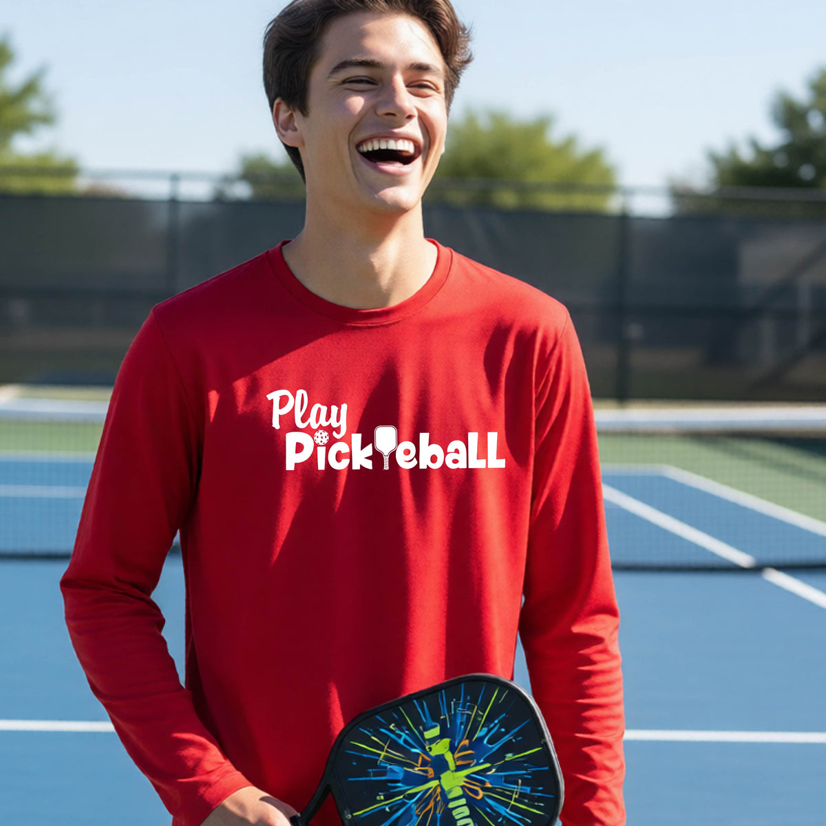 A young person on a pickleball court wearing a red long-sleeve t-shirt with a white graphic of the text 'PLAY PICKLEBALL' across the front.