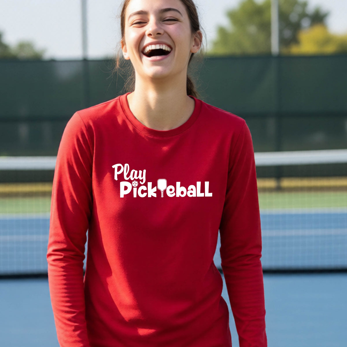 A young person on a pickleball court wearing a red long-sleeve t-shirt with a white graphic of the text 'PLAY PICKLEBALL' across the front.