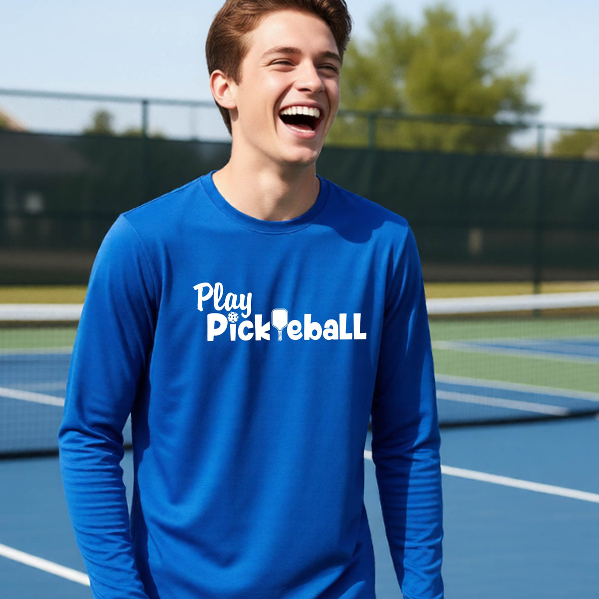 A young person on a pickleball court wearing a royal blue long-sleeve t-shirt with a white graphic of the text 'PLAY PICKLEBALL' across the front.