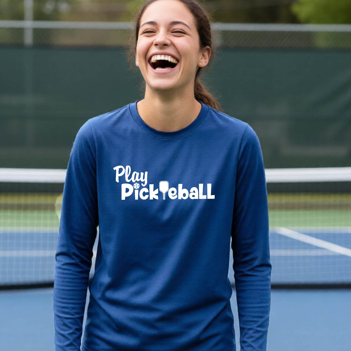 A young person on a pickleball court wearing a royal blue long-sleeve t-shirt with a white graphic of the text 'PLAY PICKLEBALL' across the front.