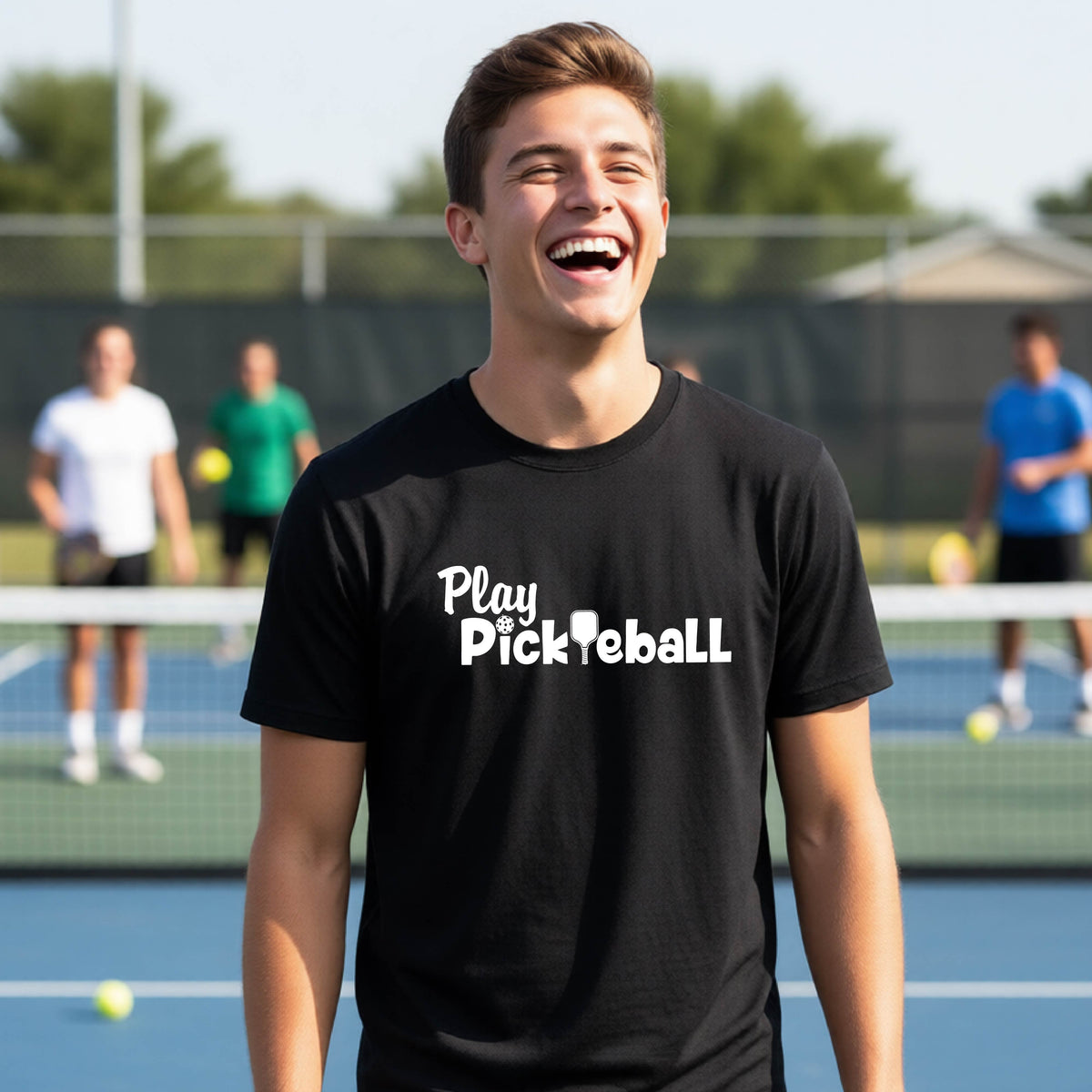 A young person laughing on an outdoor pickleball court wearing a black t-shirt with the white text 'Play Pickleball' printed on the chest.