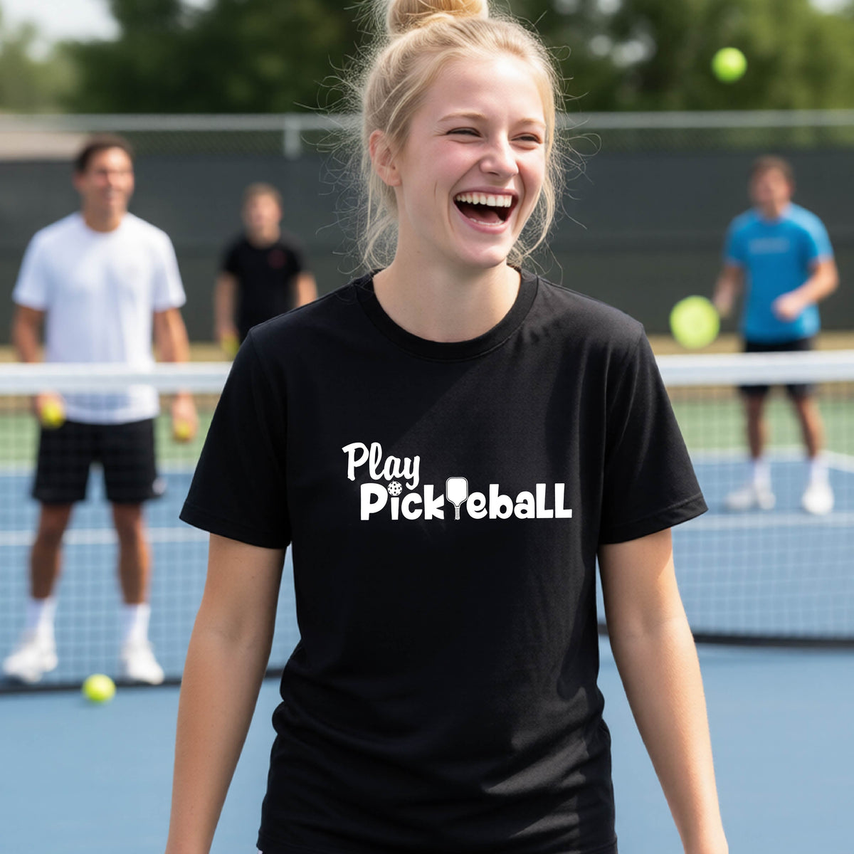 A young person laughing on an outdoor pickleball court wearing a black t-shirt with the white text 'Play Pickleball' printed on the chest.