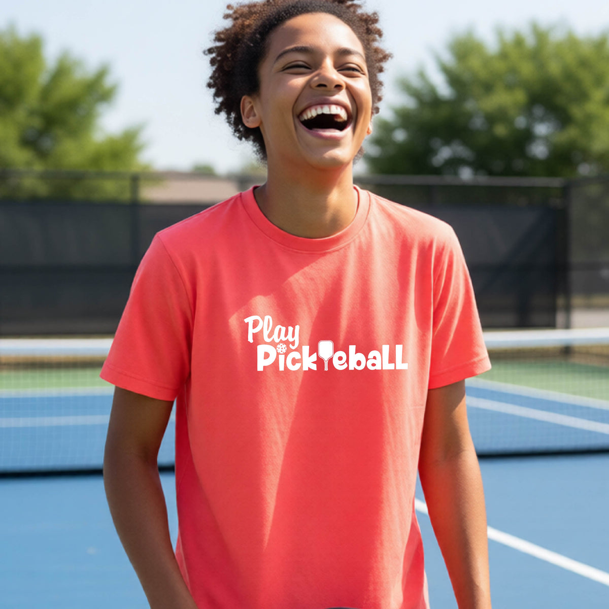 A young person laughing on an outdoor pickleball court wearing a bright coral pink t-shirt with the white text 'Play Pickleball' printed on the chest.
