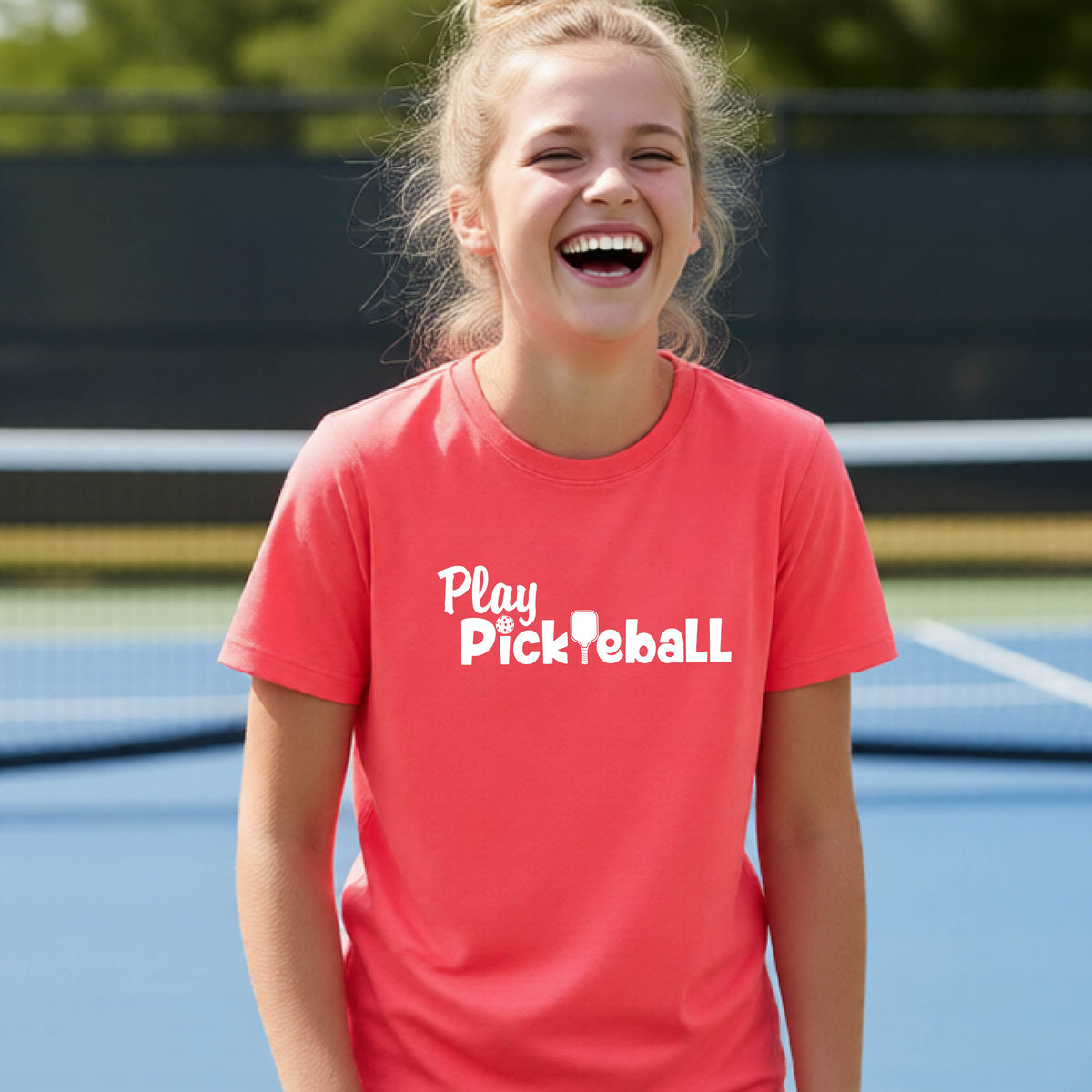 A young person laughing on an outdoor pickleball court wearing a bright coral pink t-shirt with the white text 'Play Pickleball' printed on the chest.