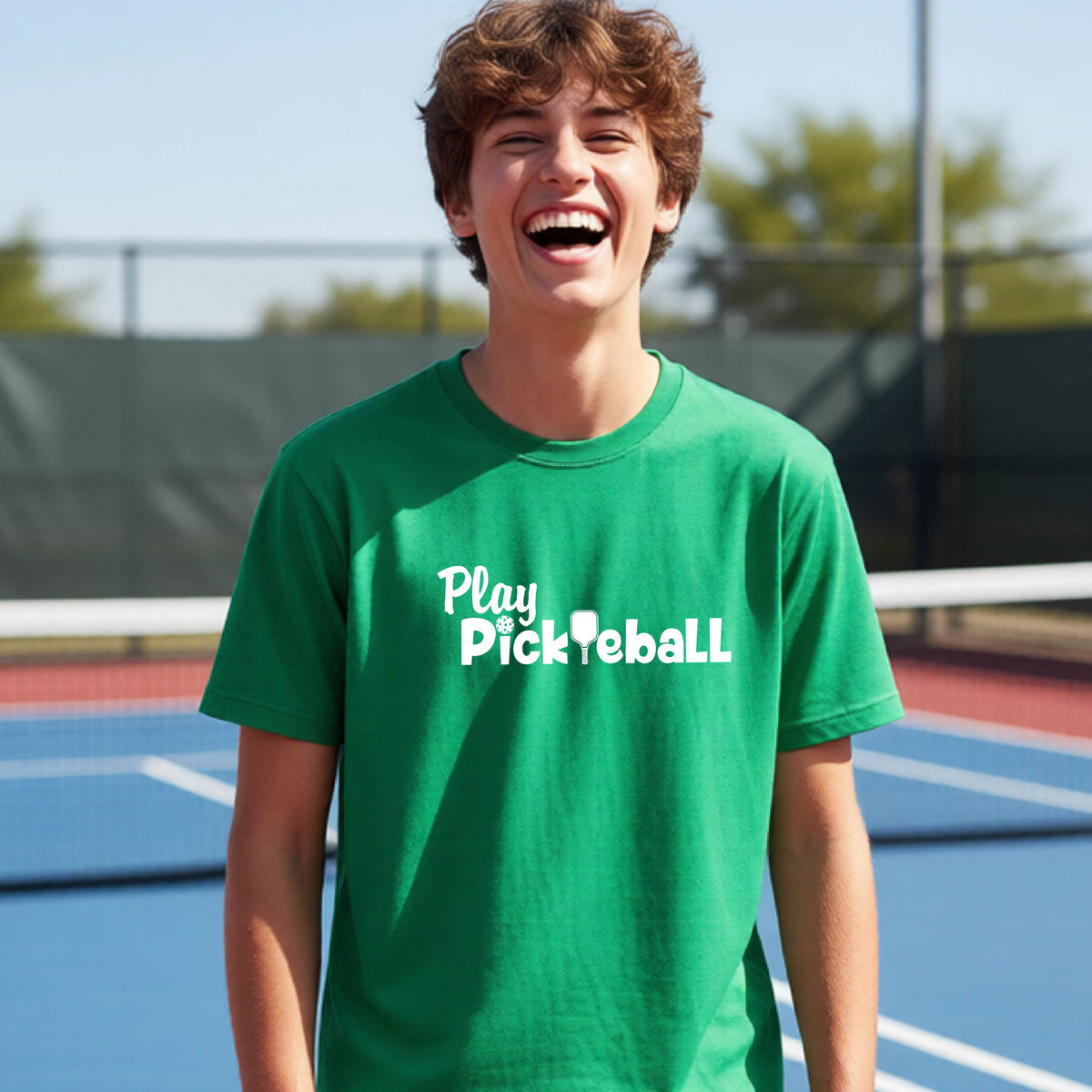 A young person laughing on an outdoor pickleball court wearing a bright green t-shirt with the white text 'Play Pickleball' printed on the chest.