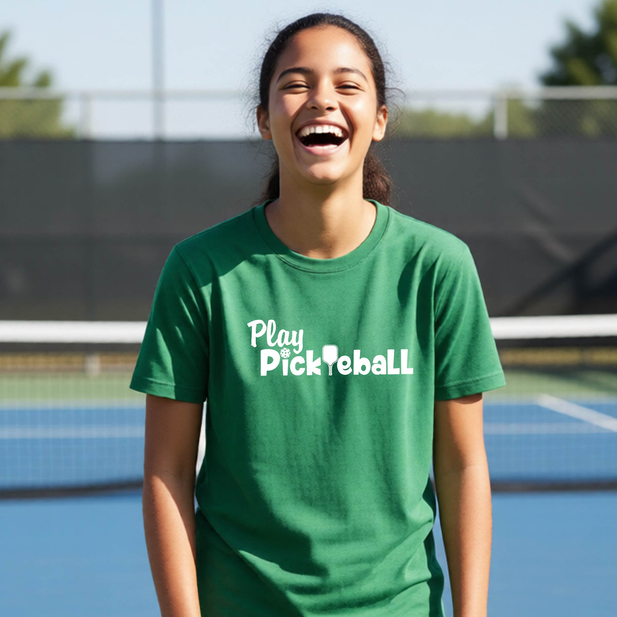 A young person laughing on an outdoor pickleball court wearing a green t-shirt with the white text 'Play Pickleball' printed on the chest.