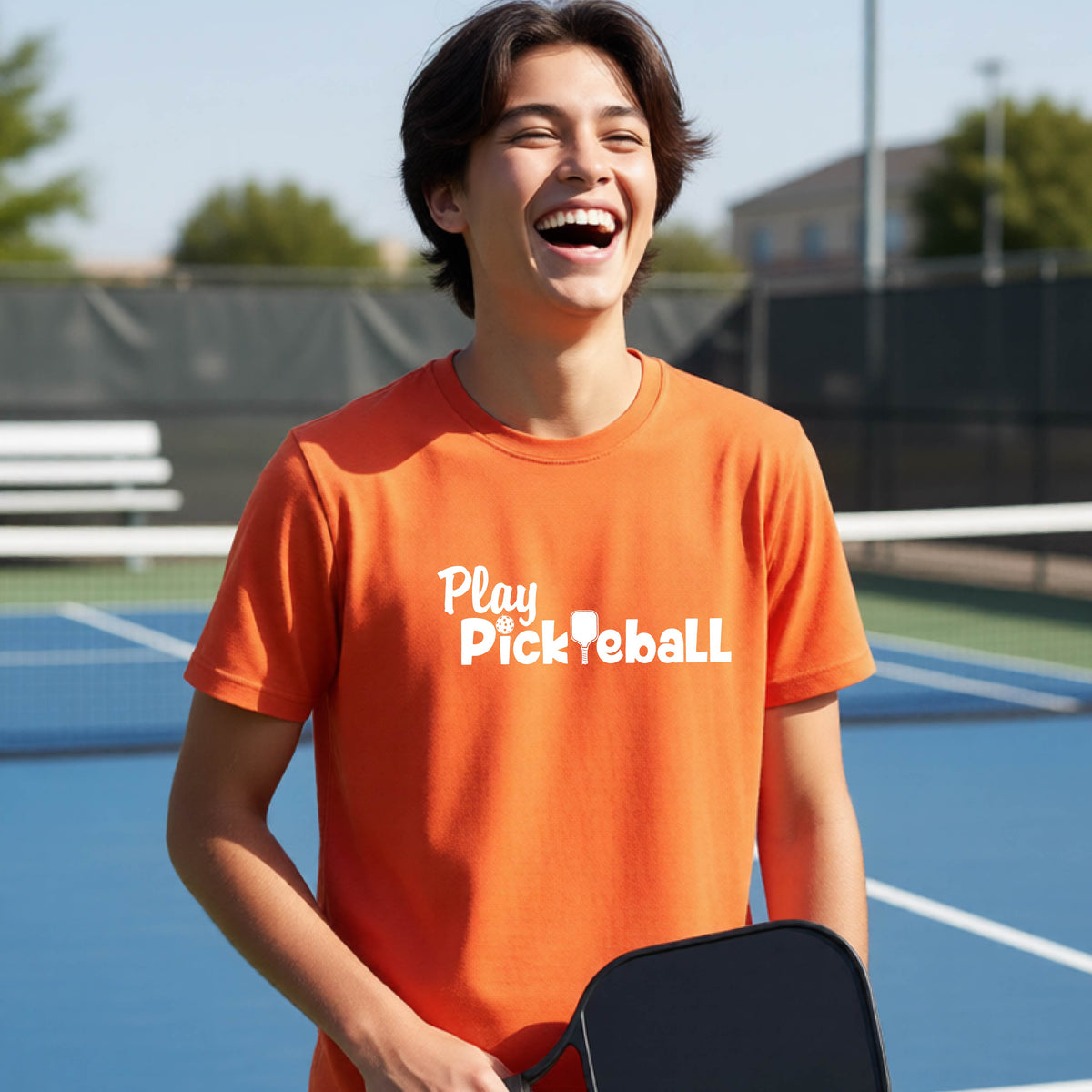 A young person laughing on an outdoor pickleball court wearing a bright orange t-shirt with the white text 'Play Pickleball' printed on the chest.