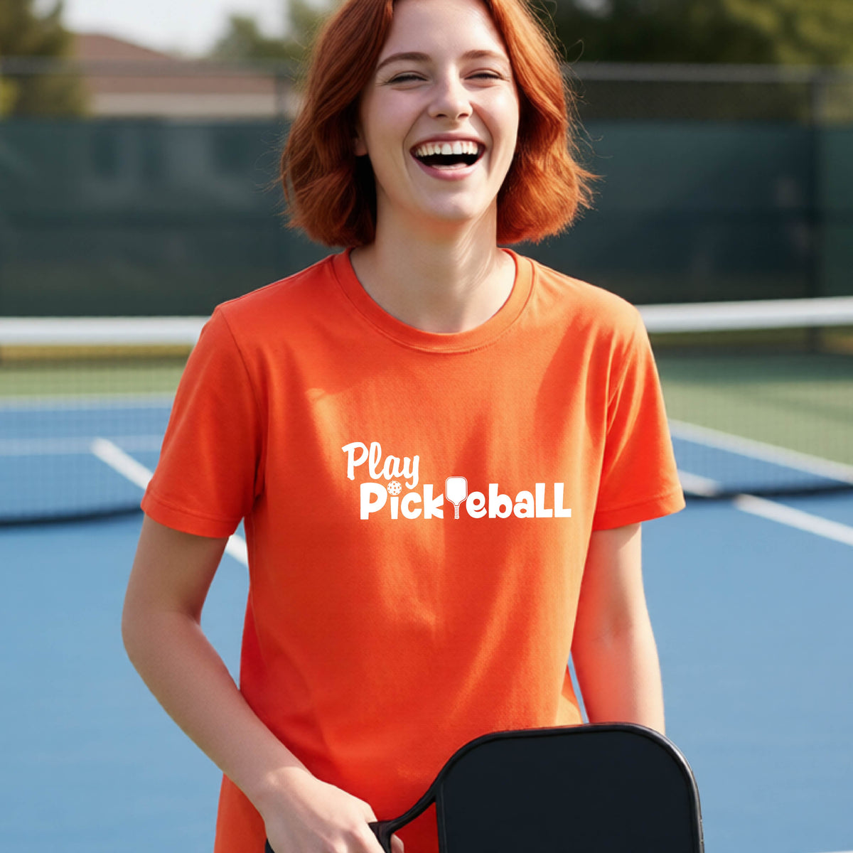 A young person laughing on an outdoor pickleball court wearing a bright orange t-shirt with the white text 'Play Pickleball' printed on the chest.