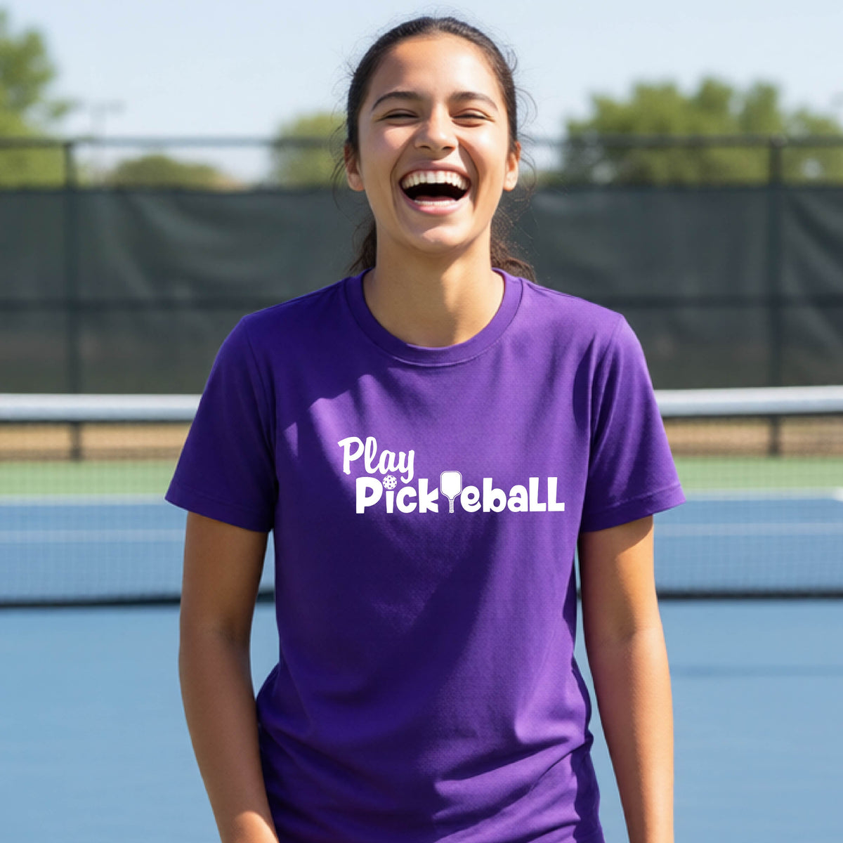 A young person laughing on an outdoor pickleball court wearing a purple t-shirt with the white text 'Play Pickleball' printed on the front. 