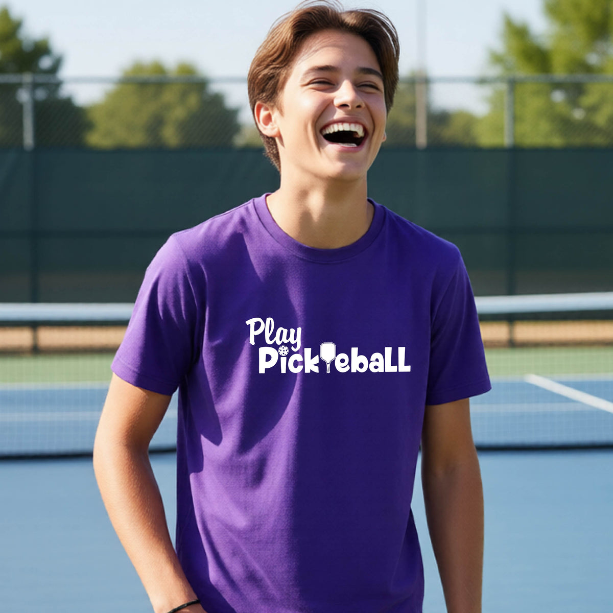 A young person laughing on an outdoor pickleball court wearing a purple t-shirt with the white text 'Play Pickleball' printed on the chest.