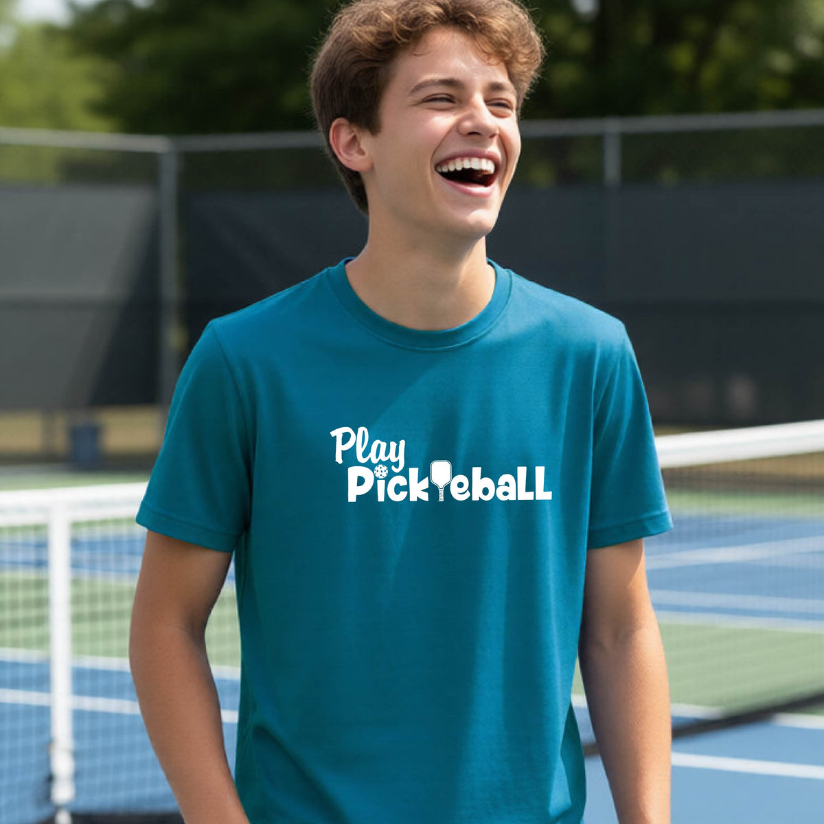 A young person laughing on an outdoor pickleball court wearing a teal t-shirt with the white text 'Play Pickleball' printed on the chest.