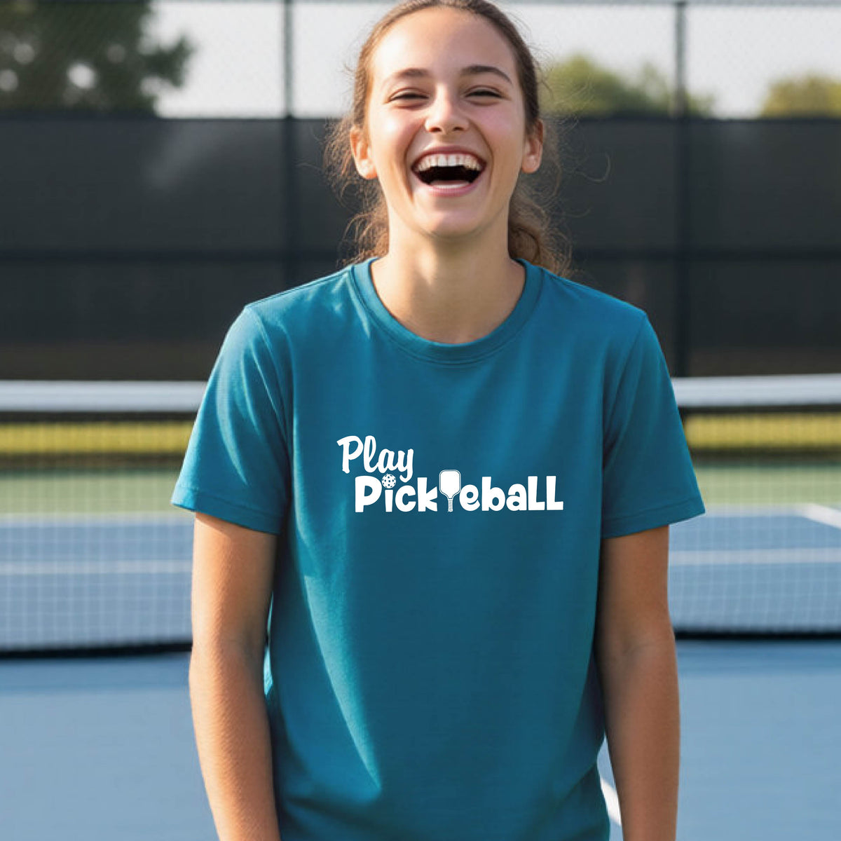A young person laughing on an outdoor pickleball court wearing a teal t-shirt with the white text 'Play Pickleball' printed on the chest.