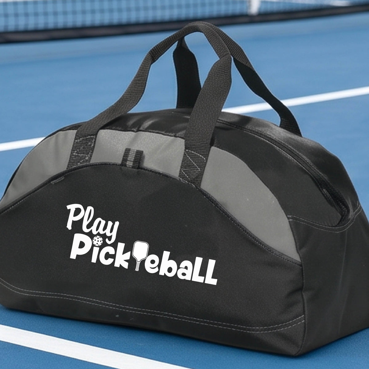 A duffel bag for pickleball on a green picleball court. The bag is black with gray panels, featuring the text 'Play Pickleball' in white on the front.