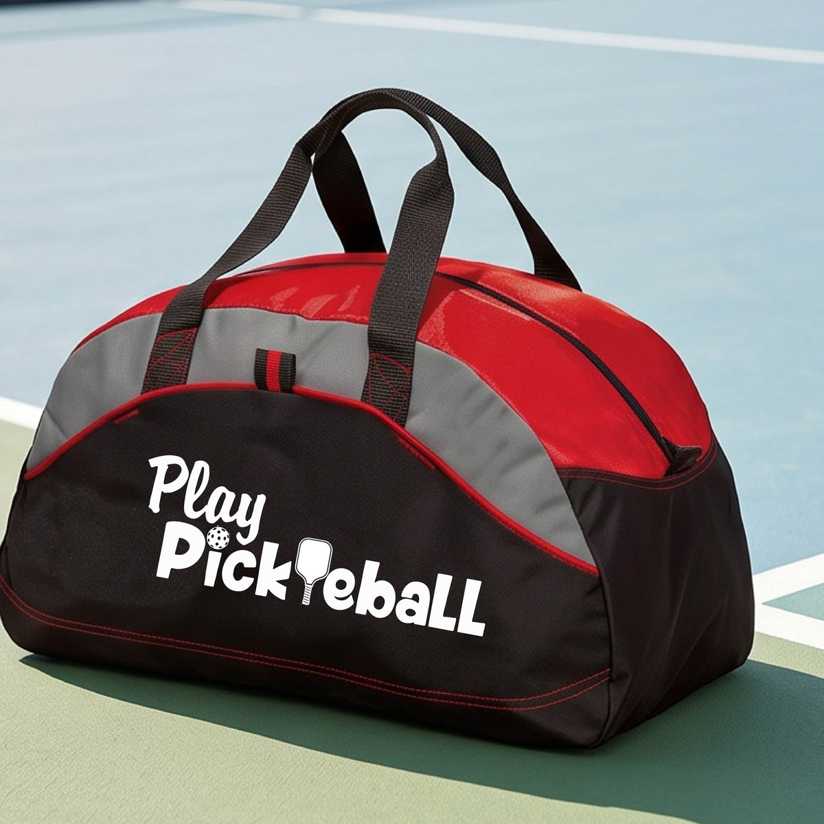 A duffel bag for pickleball on a green picleball court. The bag is red, black and lime green with gray panels, featuring the text 'Play Pickleball' in white on the front.