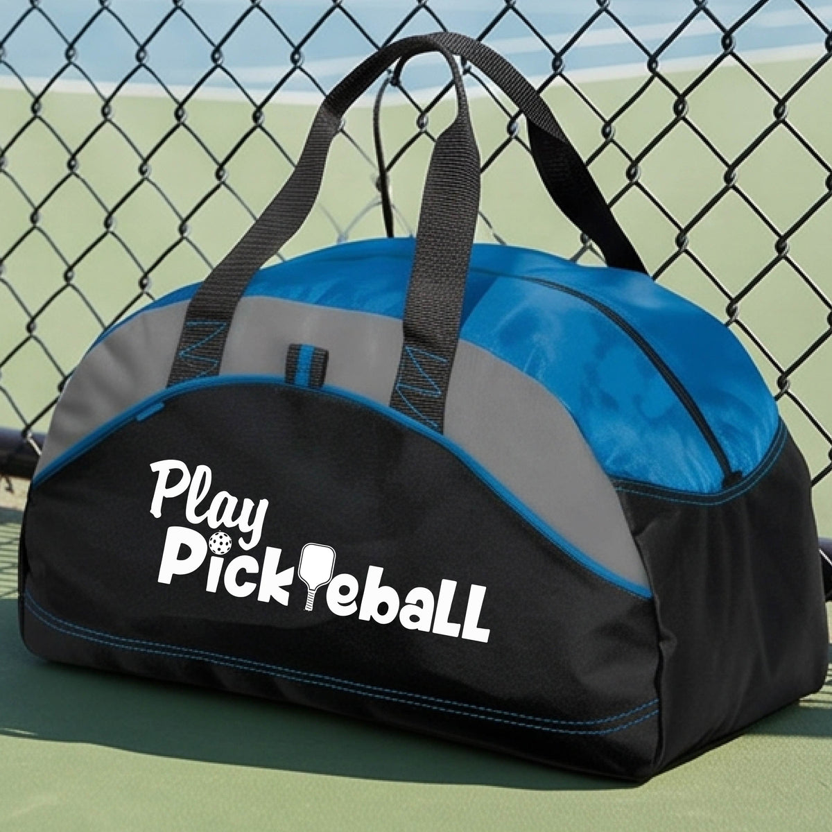 A duffel bag for pickleball on a green picleball court. The bag is black and royal blue with gray panels, featuring the text 'Play Pickleball' in white on the front.