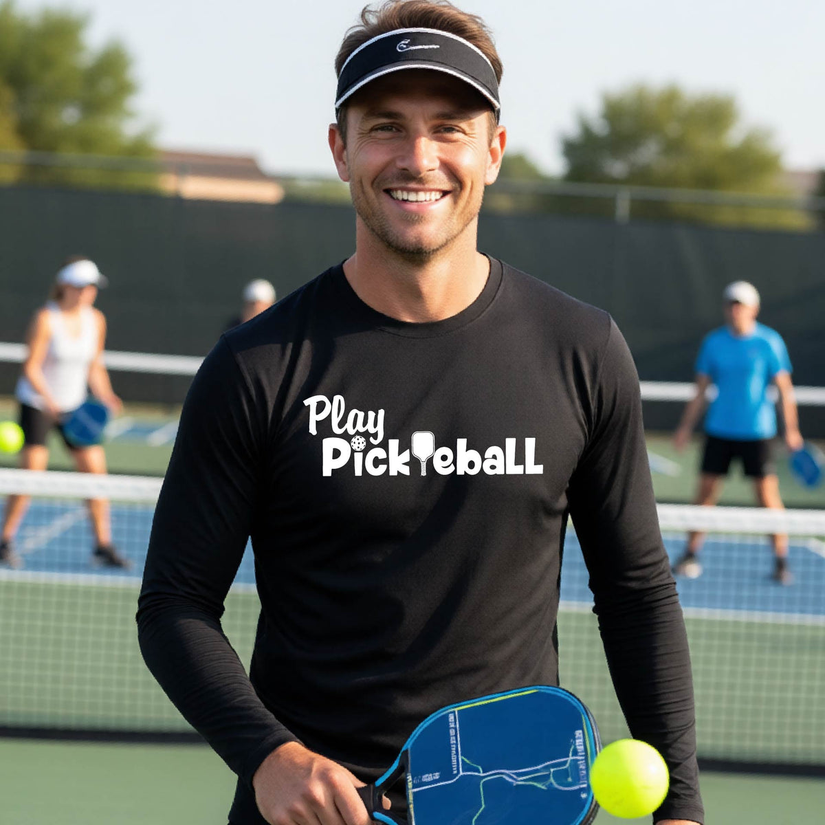 A smiling man on a pickleball court wearing a black long-sleeve shirt with 'Play Pickleball' printed in white across the chest.