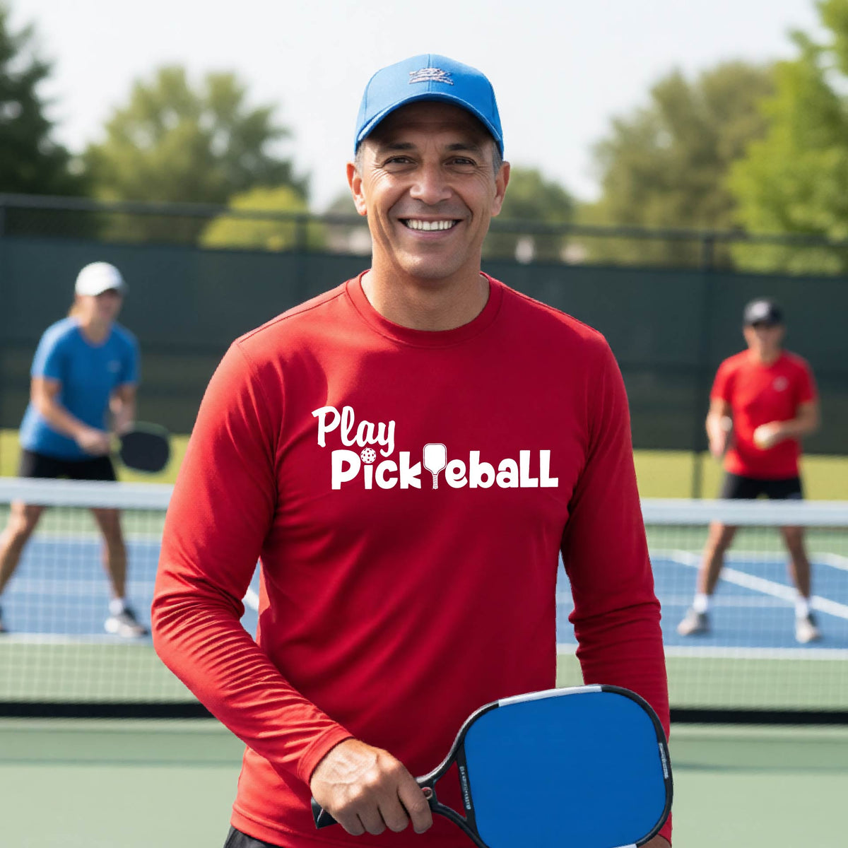A smiling man on a pickleball court wearing a red long-sleeve shirt with 'Play Pickleball' printed in white across the chest.