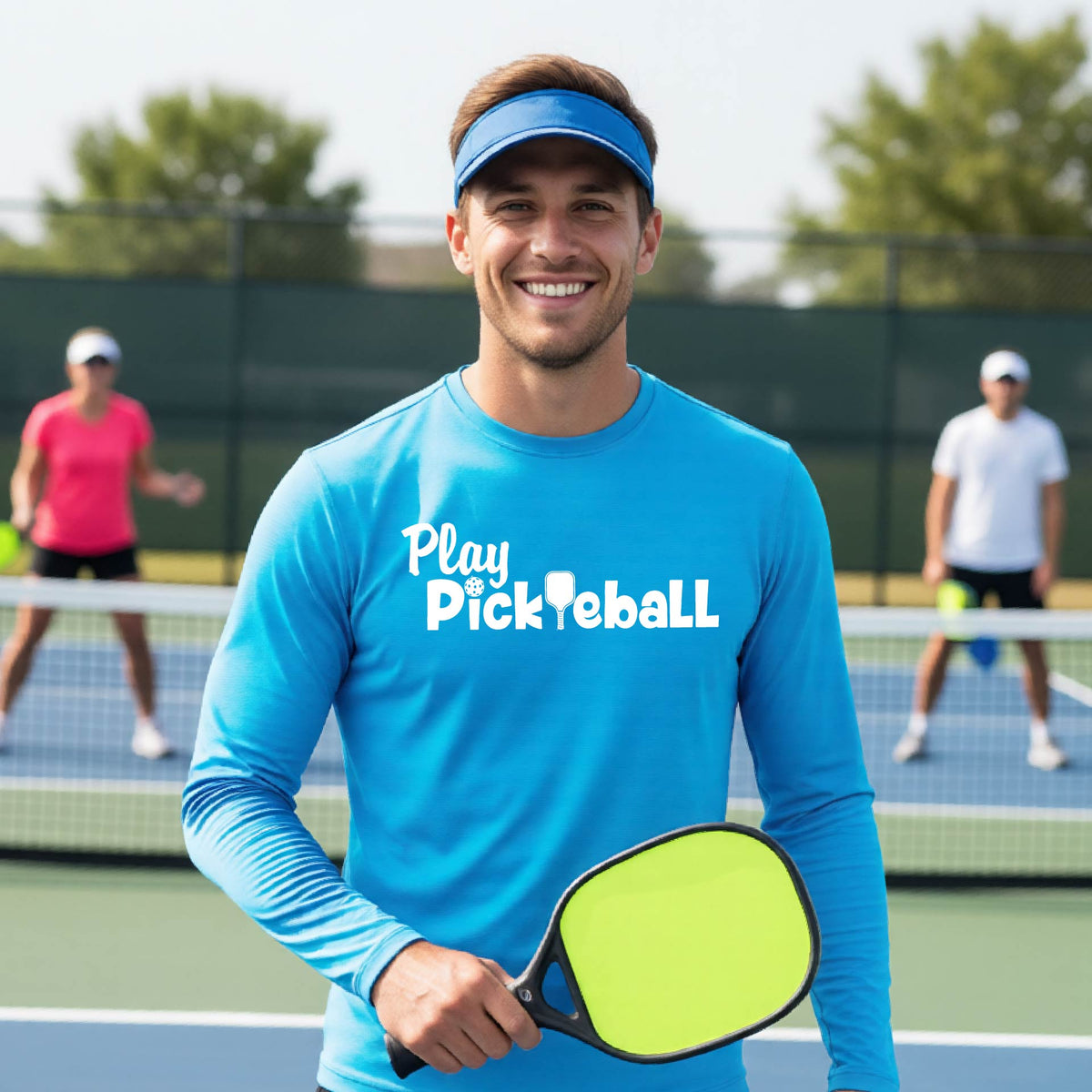 A smiling man on a pickleball court wearing a bright blue long-sleeve shirt with 'Play Pickleball' printed in white across the chest.