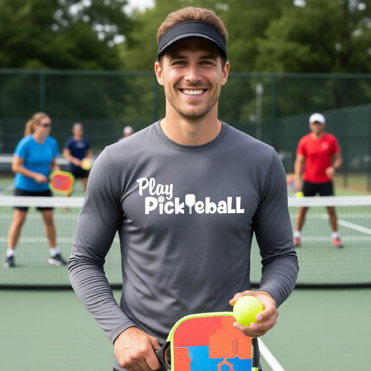 A smiling man on a pickleball court wearing a dark gray long-sleeve shirt with 'Play Pickleball' printed in white across the chest.