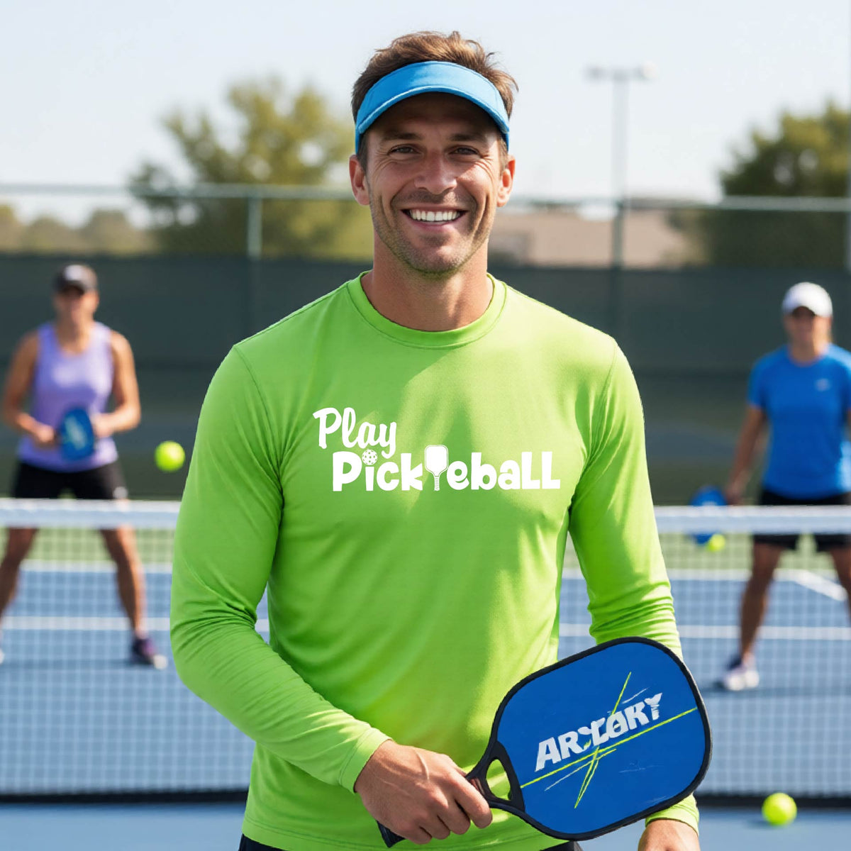 A smiling man on a pickleball court wearing a lime green long-sleeve shirt with 'Play Pickleball' printed in white across the chest.