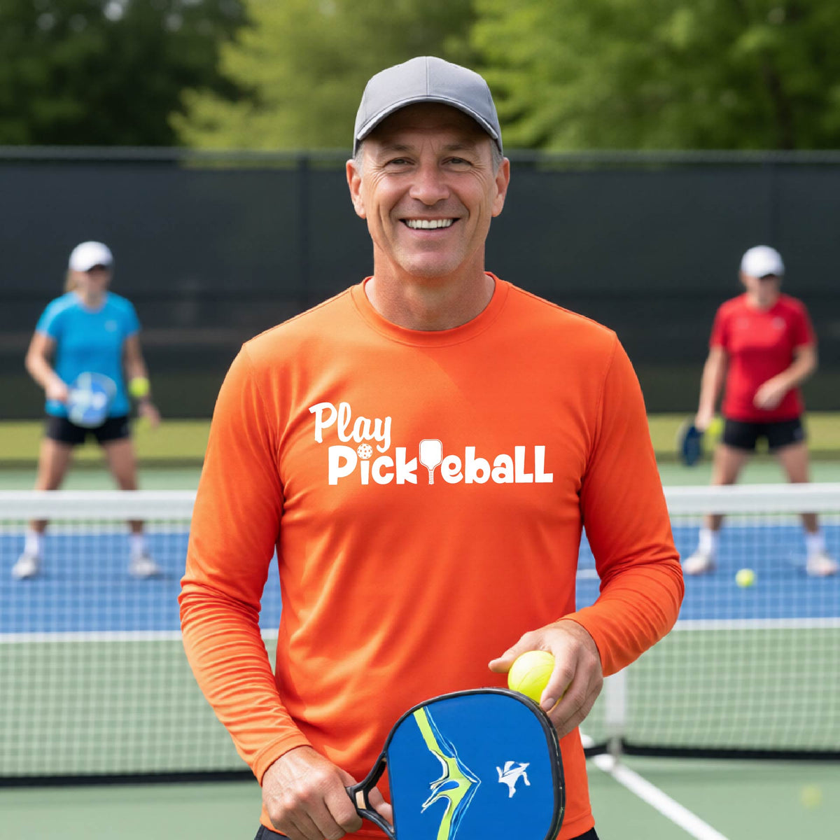 A smiling man on a pickleball court wearing a bright orange long-sleeve shirt with 'Play Pickleball' printed in white across the chest.
