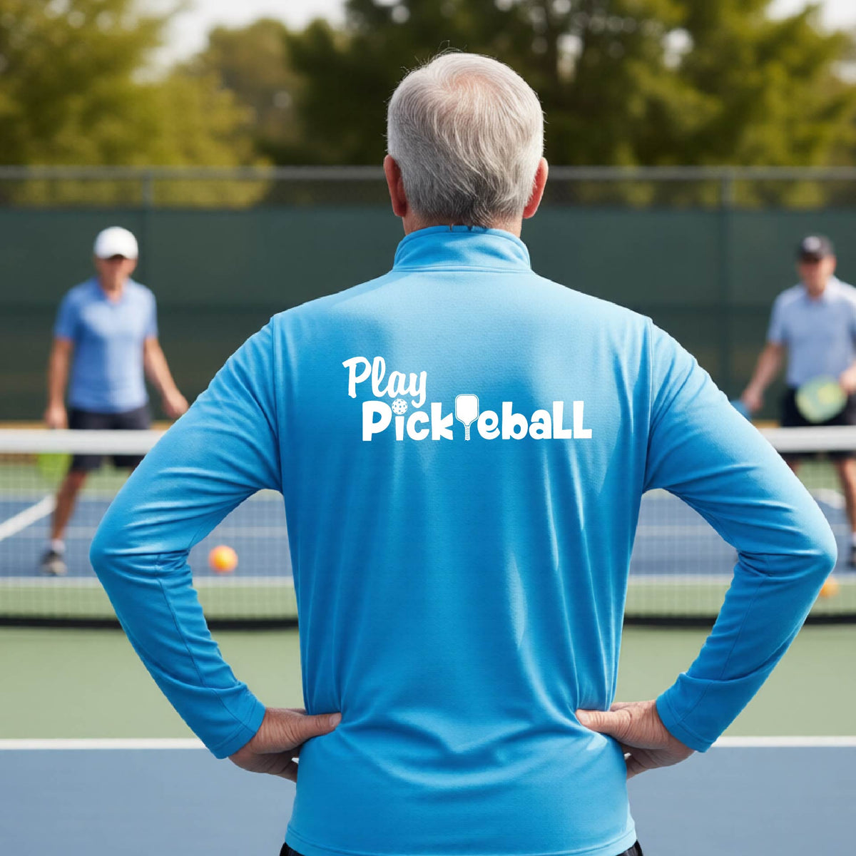 A man viewed from behind on a pickleball court, wearing a bright blue long-sleeve shirt with the white text 'Play Pickleball' printed across the back. 