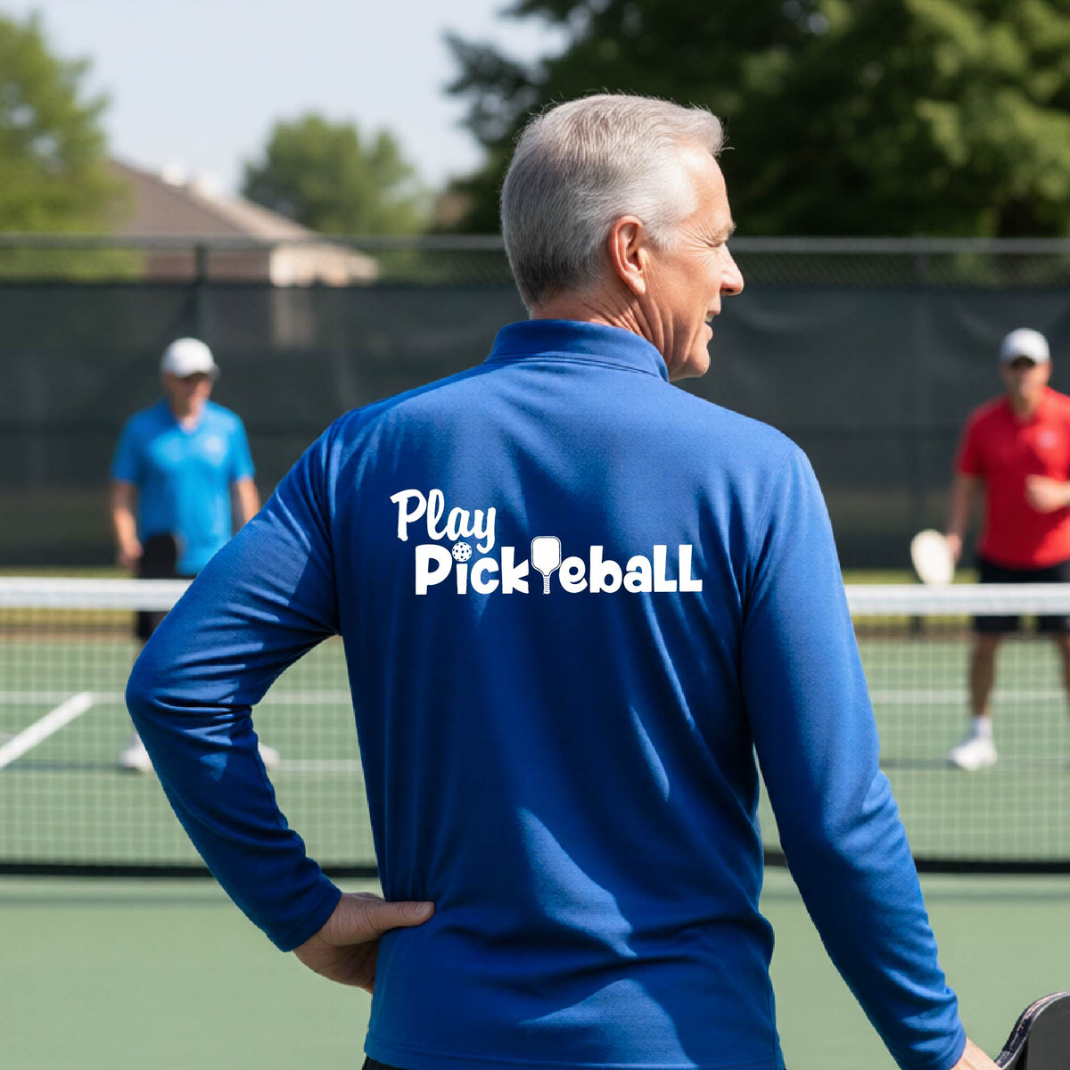 A man viewed from behind on a pickleball court, wearing a royal blue long-sleeve shirt with the white text 'Play Pickleball' printed across the back. 