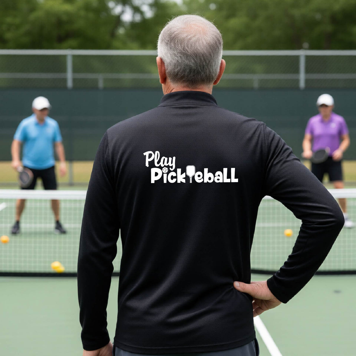 A man viewed from behind on a pickleball court, wearing a black long-sleeve shirt with the white text 'Play Pickleball' printed across the back. 