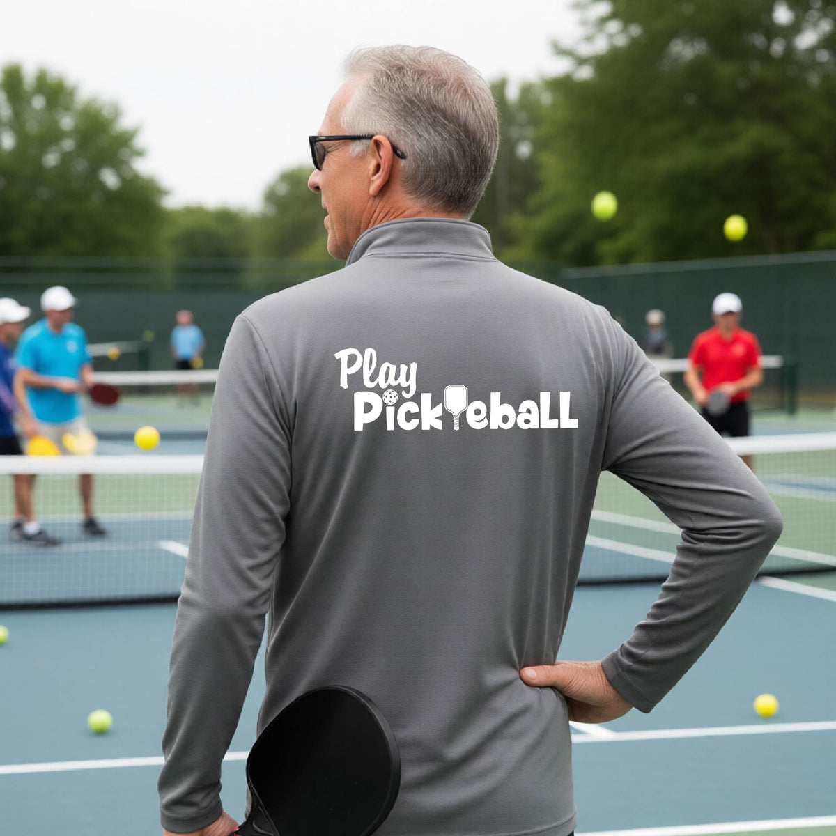 A man viewed from behind on a pickleball court, wearing a gray long-sleeve shirt with the white text 'Play Pickleball' printed across the back. 