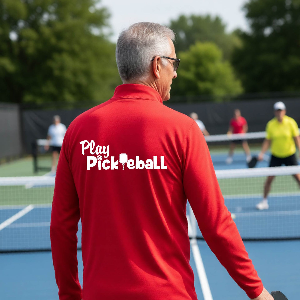 A man viewed from behind on a pickleball court, wearing a red long-sleeve shirt with the white text 'Play Pickleball' printed across the back. 