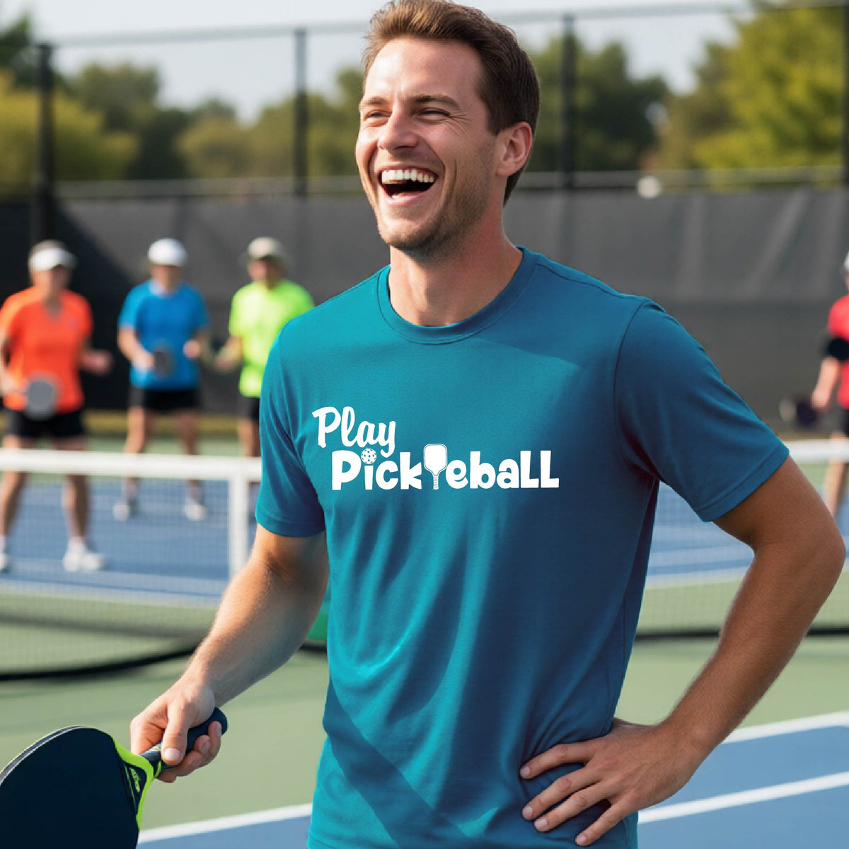 A man laughing on a pickleball court wearing a teal t-shirt with 'Play Pickleball' printed in white across the chest, holding a paddle. Other players are visible in the background on the court.