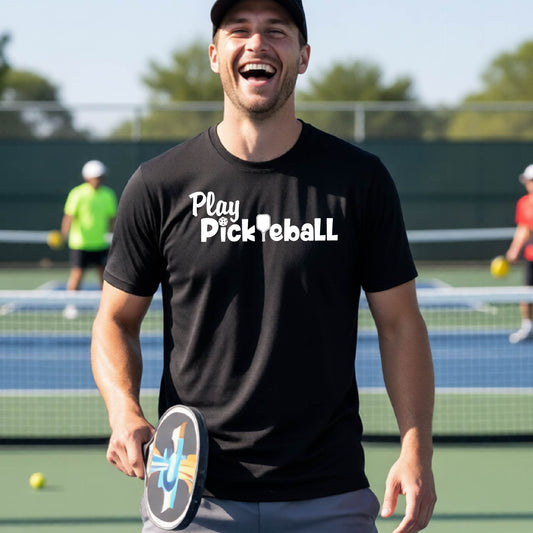 A man laughing on a pickleball court wearing a black t-shirt with the text 'Play Pickleball' in white. He is holding a pickleball paddle, with other players and a court visible in the background.