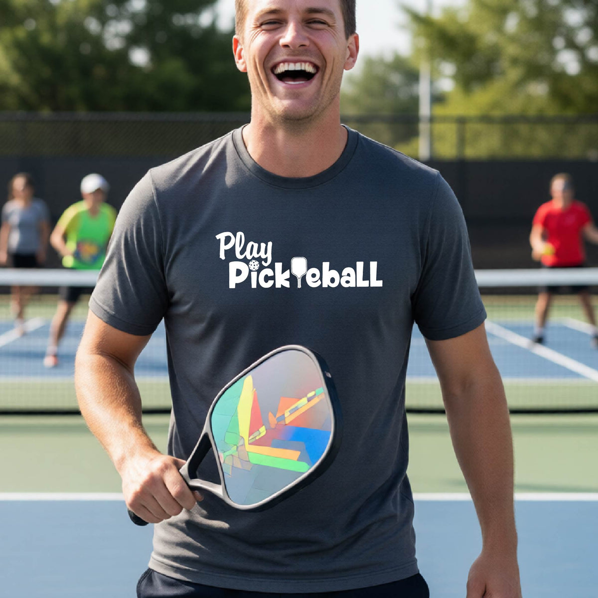 A man on a pickleball court wearing a dark gray t-shirt with the text 'Play Pickleball' in white, holding a pickleball paddle. The background shows a blurred outdoor pickleball court with other players.