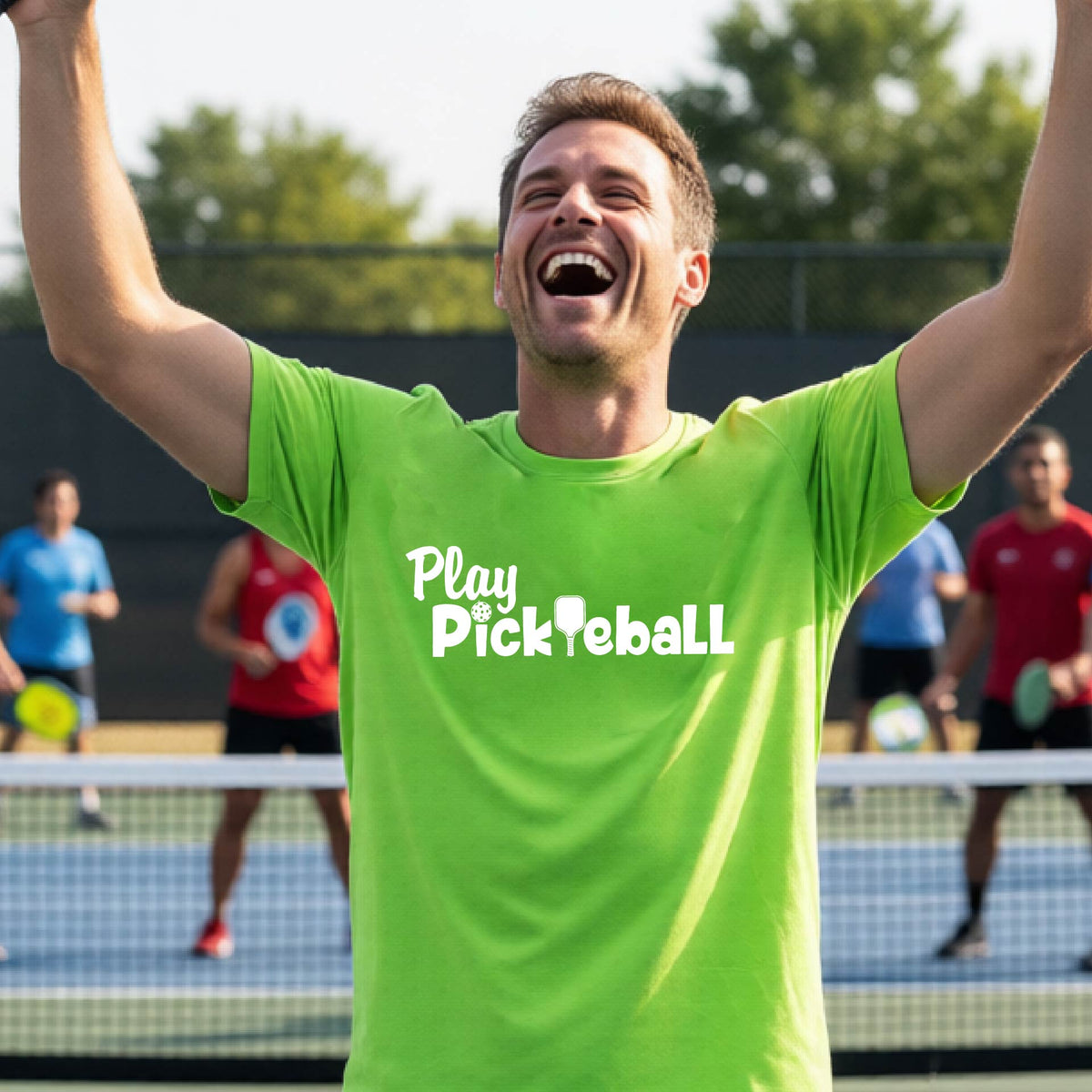 A man celebrating on a pickleball court wearing a bright lime green t-shirt with the white text 'Play Pickleball' printed on the chest. Other players are visible in the background on the court.