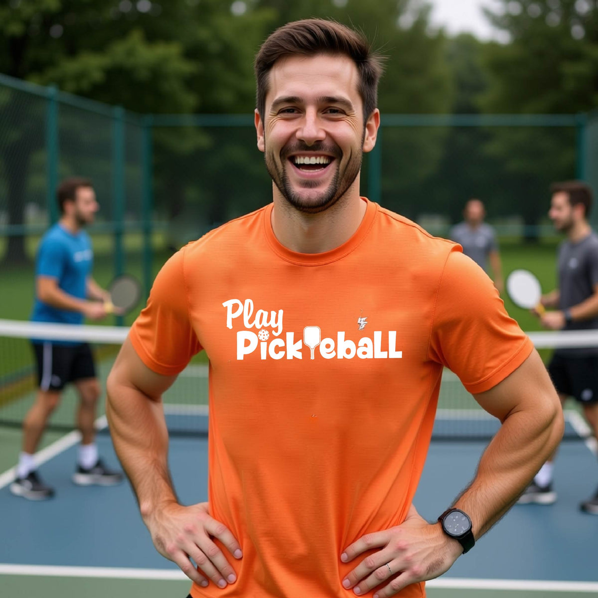 A smiling man on an outdoor tennis court wearing a bright orange t-shirt with the text 'Play Pickleball' printed in white across the chest. Other players are visible in the background on the court.