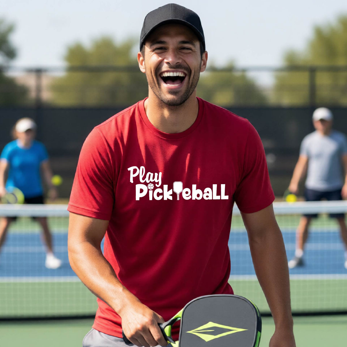 A man smiling on a pickleball court wearing a red t-shirt with 'Play Pickleball' printed in white on the chest, holding a pickleball paddle.