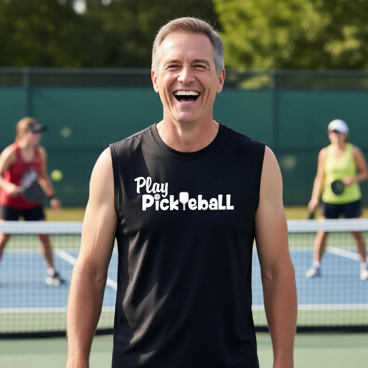 A smiling man wearing a black sleeveless t-shirt with 'Play Pickleball' printed in white on the chest, standing on a pickleball court with players blurred in the background.