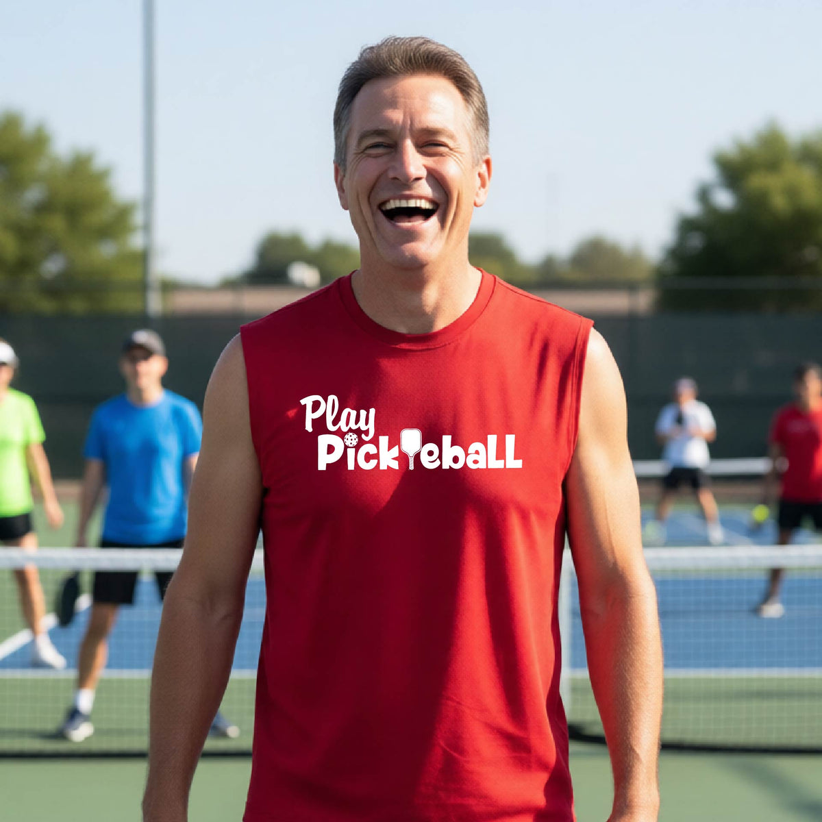 A smiling man wearing a red sleeveless t-shirt with 'Play PicklebaLL' printed in white on the chest, standing on a pickleball court with players blurred in the background.