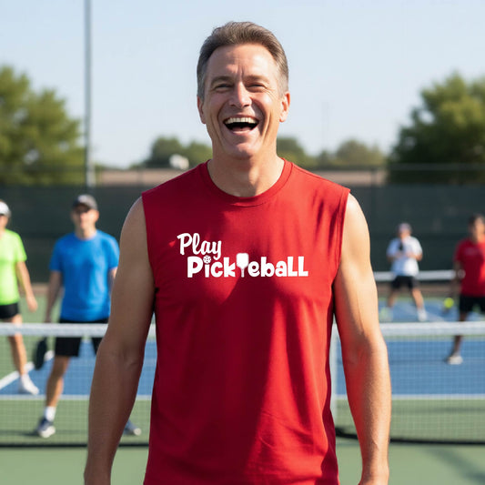 A smiling man wearing a red sleeveless t-shirt with 'Play PicklebaLL' printed in white on the chest, standing on a pickleball court with players blurred in the background.
