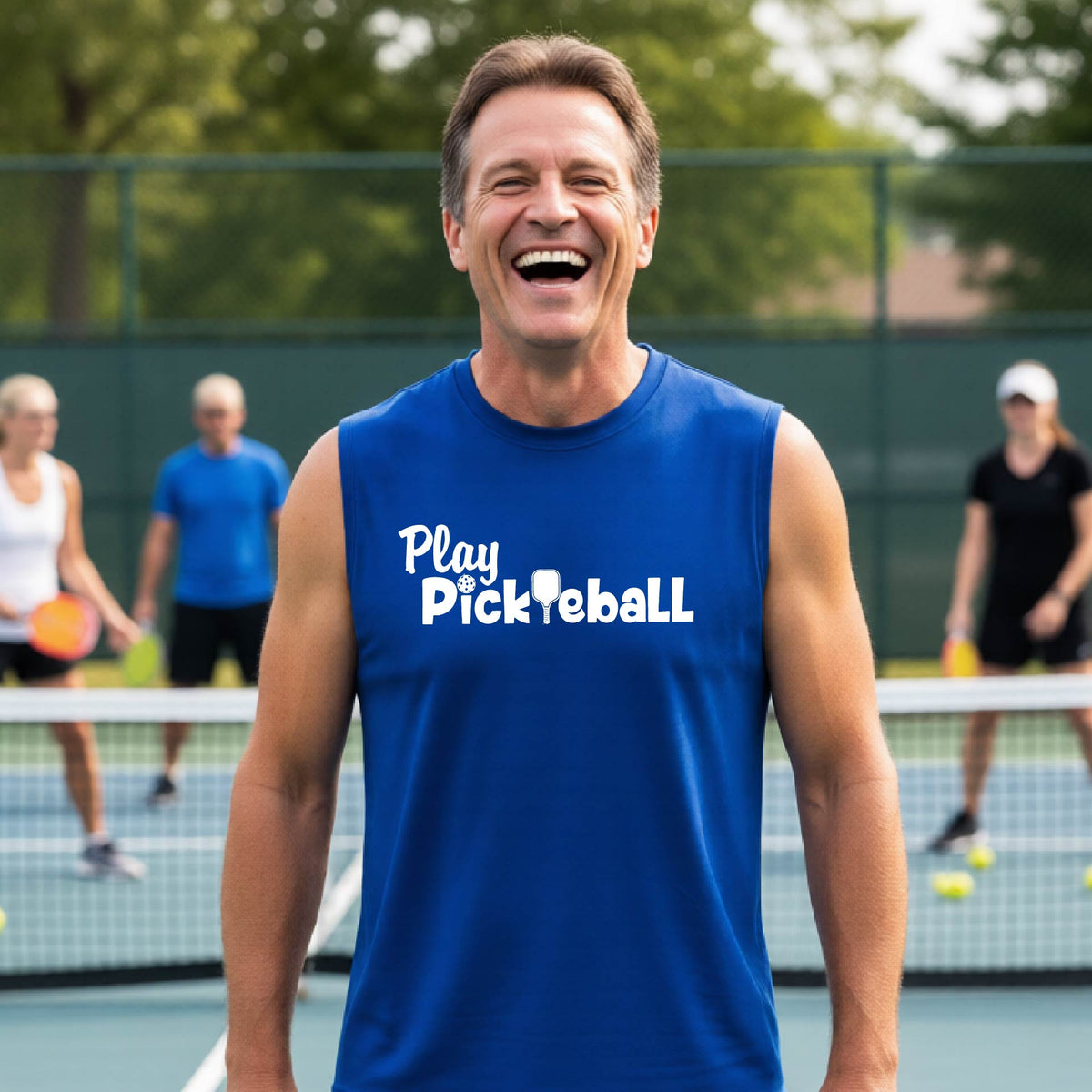 A smiling man in a blue sleeveless shirt with 'Play Pickleball' printed on the front stands on an outdoor pickleball court, with players blurred in the background.